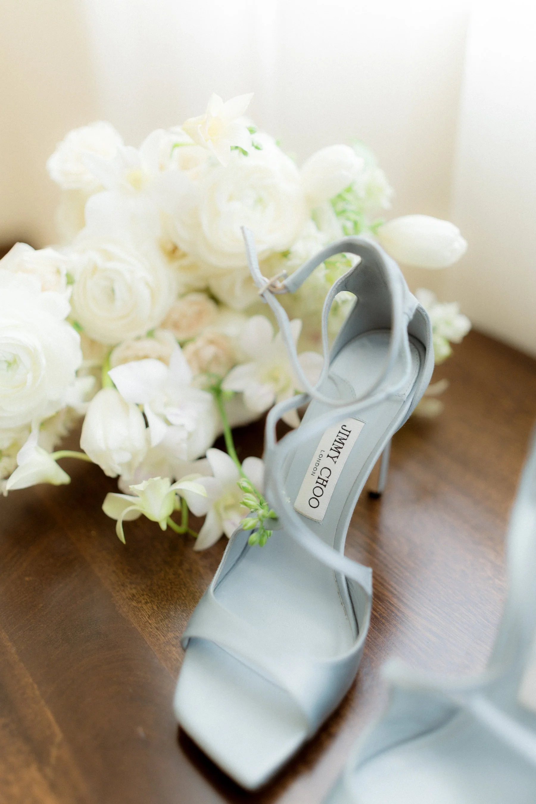 Un zapato de tacón gris de la marca JIMMY CHOO, rodeado de flores blancas en una mesa de madera.