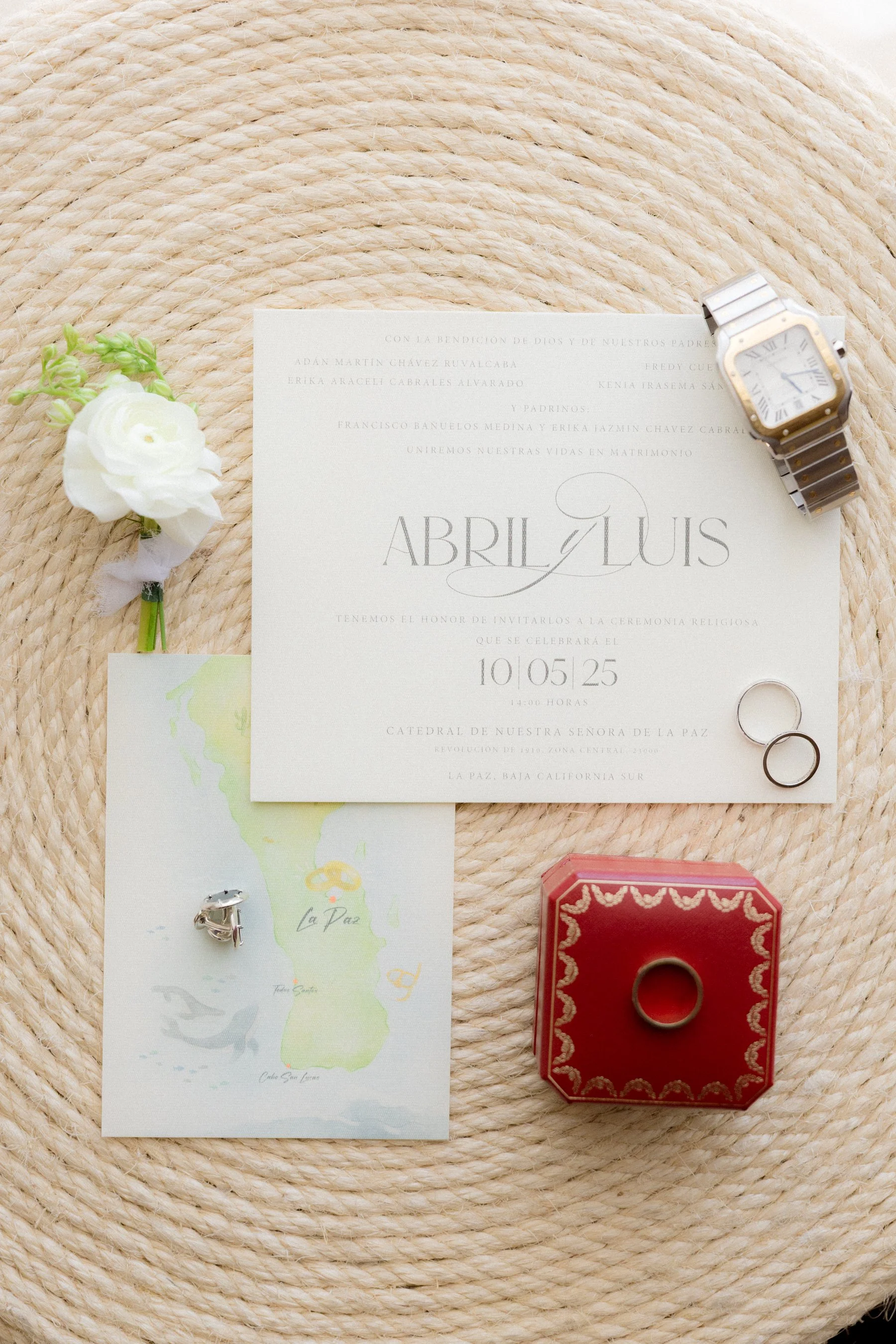Una invitación de boda que dice 'Abril y Luis', con detalles de alianzas, un reloj, una flor y un mapa de La Paz, en un fondo de tapete trenzado.