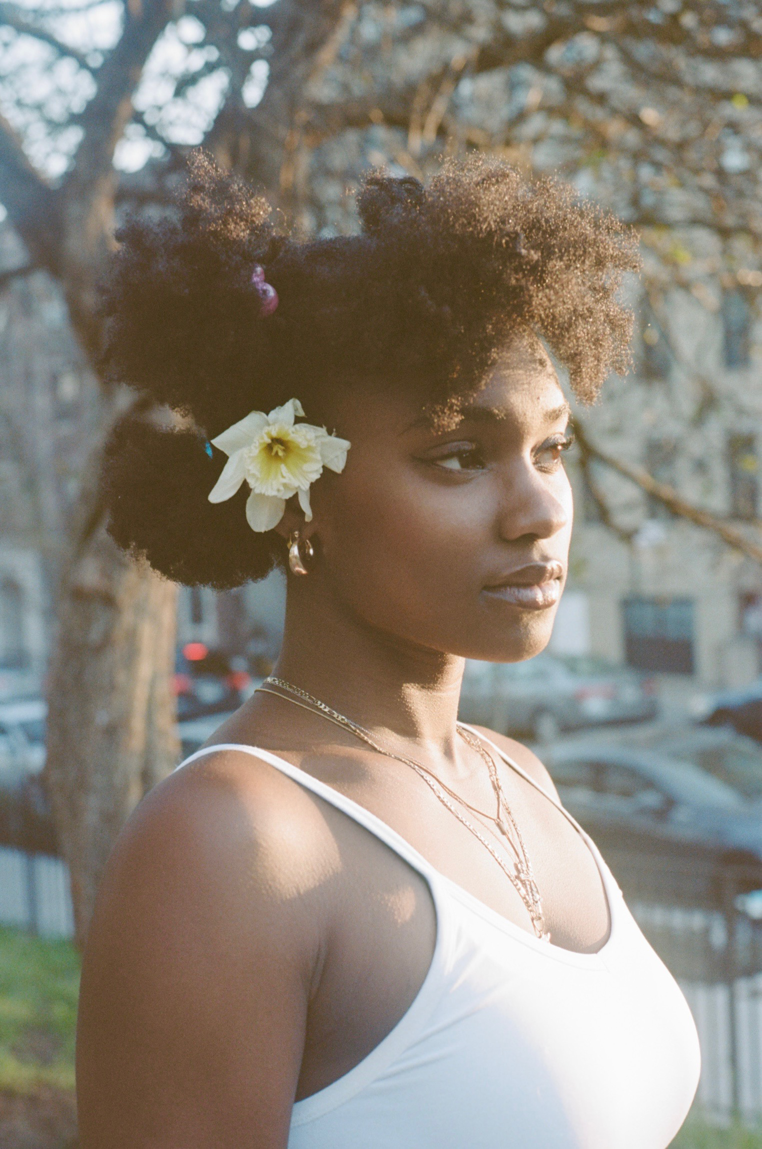 A young Black woman with natural afro hair adorned with a yellow flower wearing a white top stands outdoors during daylight, with trees and cars in the background.
