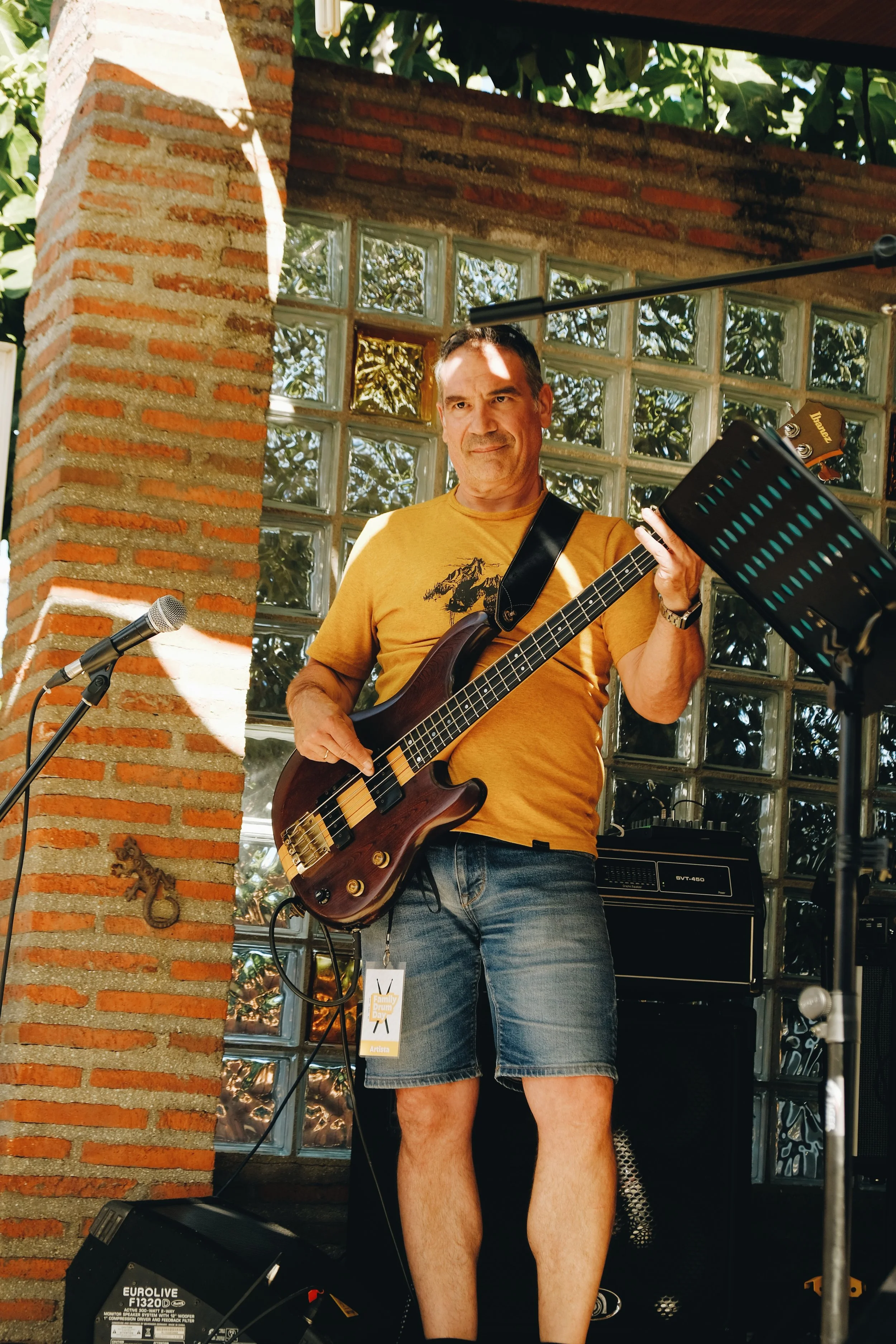 Hombre tocando una guitarra en un escenario con paredes de bloques de vidrio y ladrillos, en un entorno exterior.