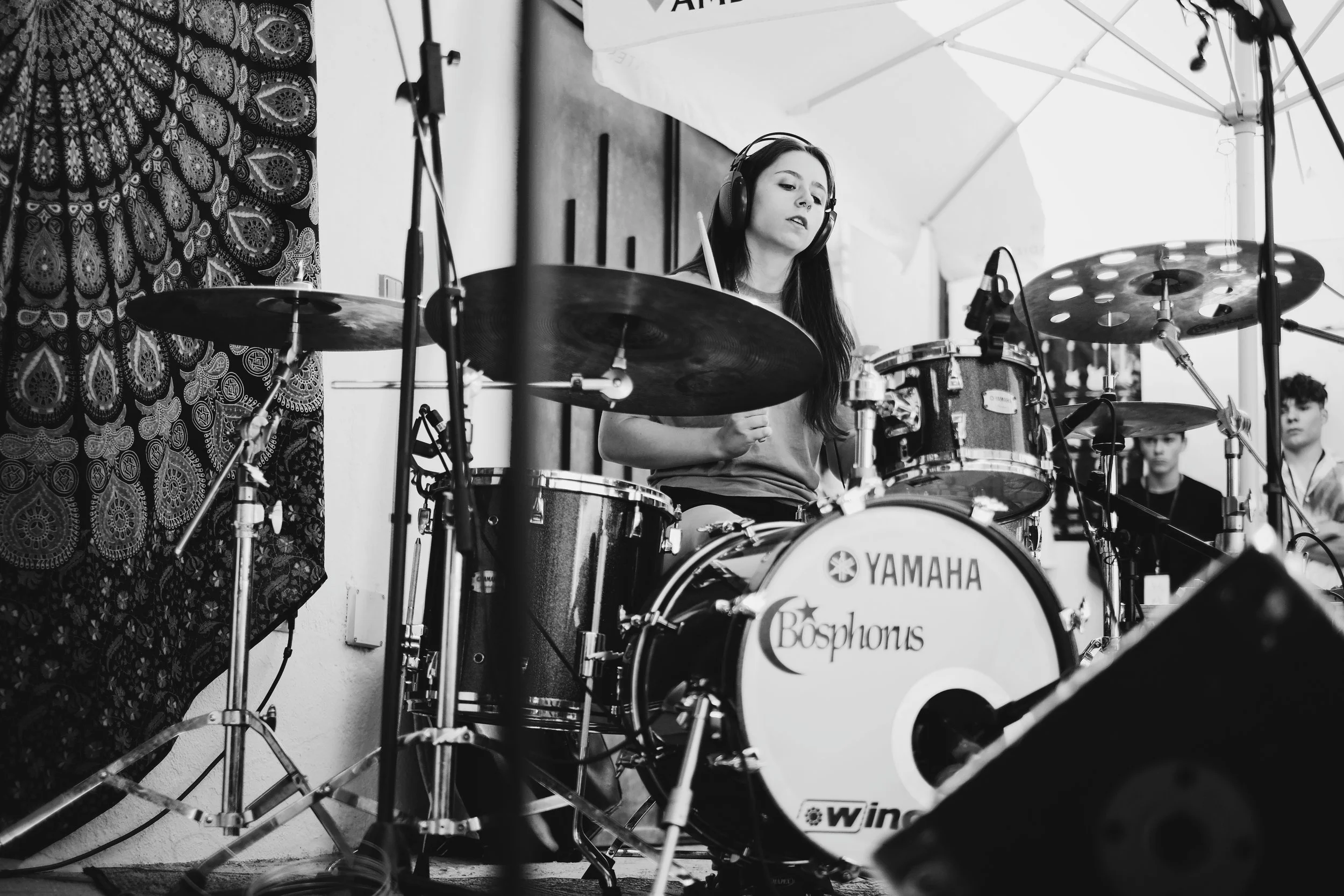 Mujer tocando batería en un escenario, en blanco y negro, con otros músicos al fondo y un paraguas en un escenario exterior.