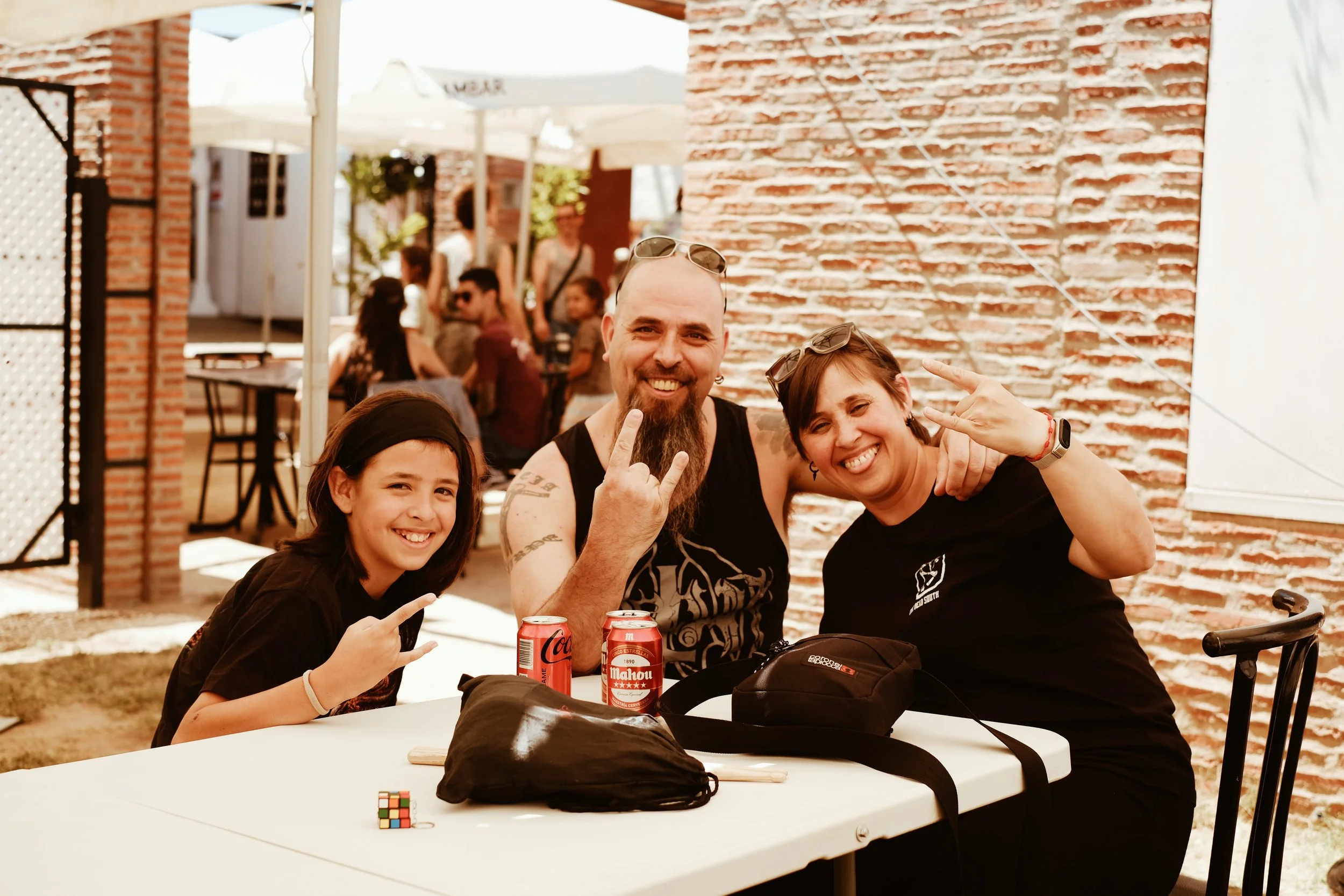 Tres personas sonrientes en una mesa al aire libre en un ambiente festivo, con una pared de ladrillos y otras personas en el fondo.