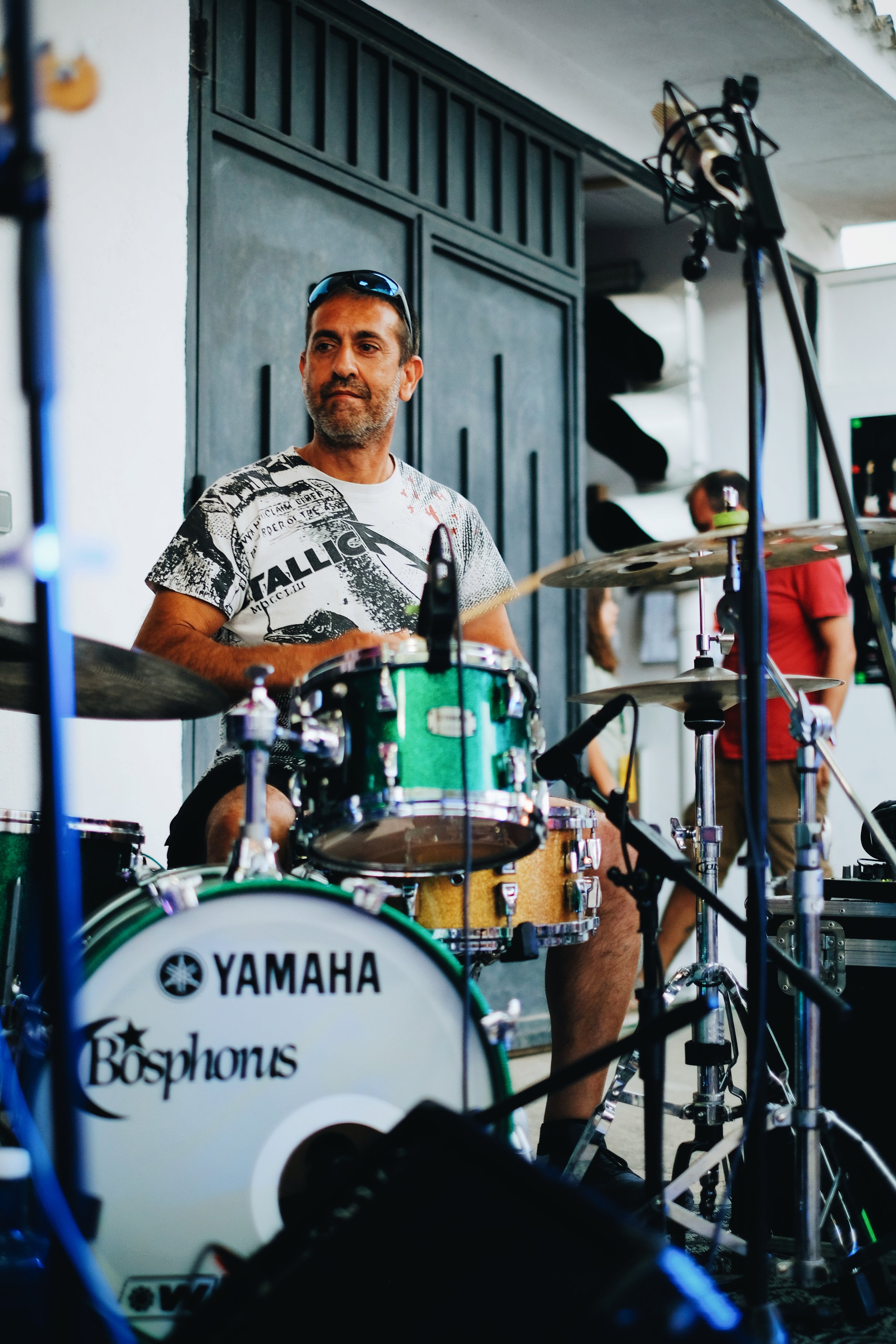 Hombre tocando la batería en un escenario, con una camiseta blanca con gráficos y gafas en la cabeza.