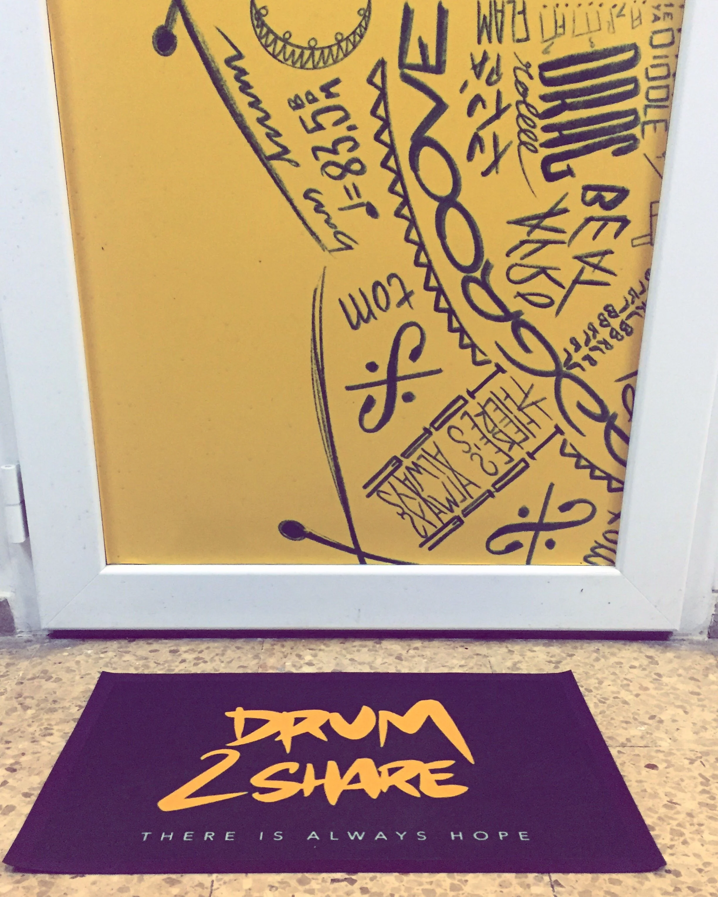 Alfombra negra con letras amarillas que dice 'DRUM 2 SHARE' y debajo 'THERE IS ALWAYS HOPE', en el suelo frente a una puerta con dibujos y frases en color negro.