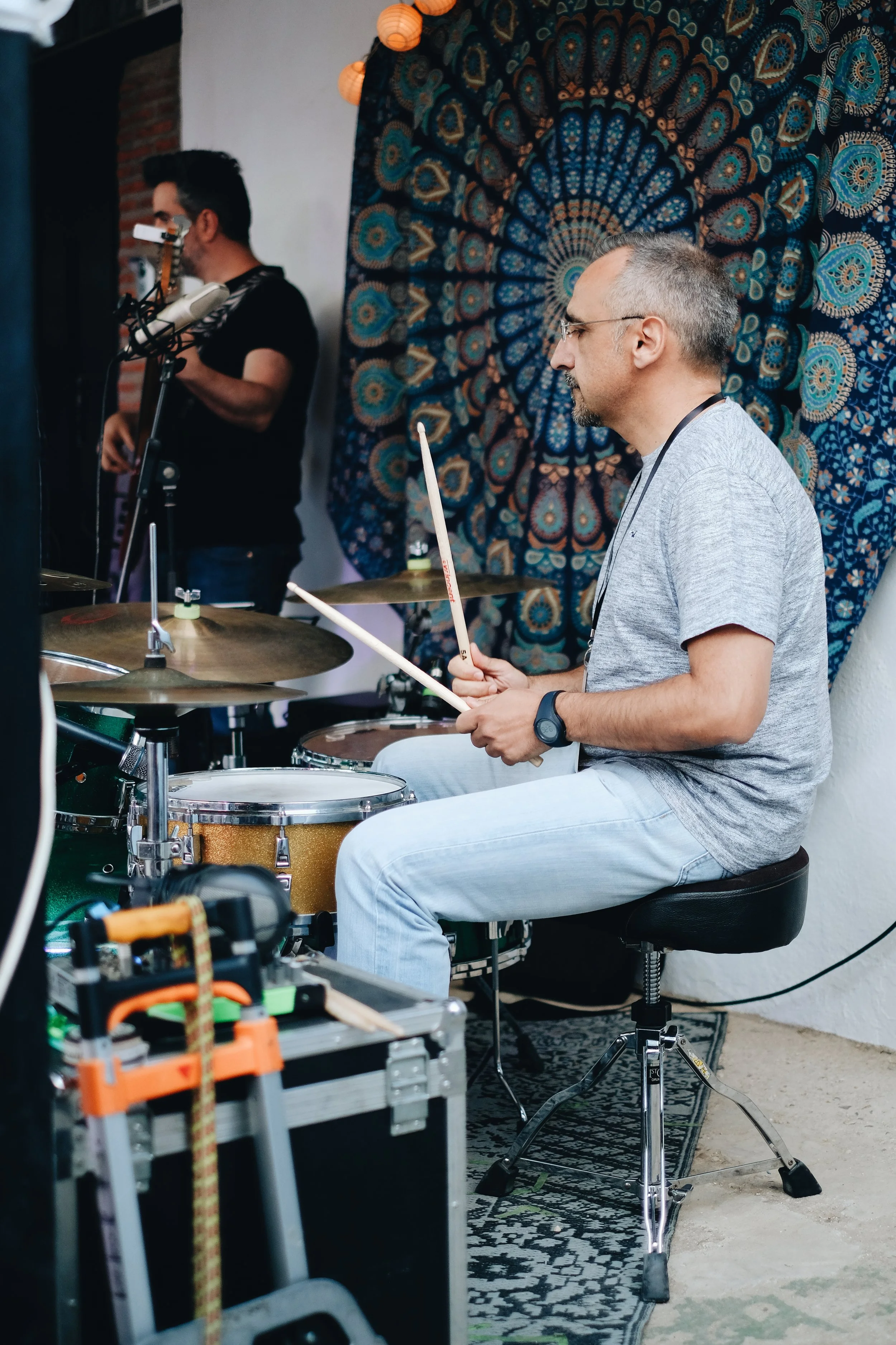 Hombre tocando la batería con una manta de mandala de fondo y otro músico en el fondo