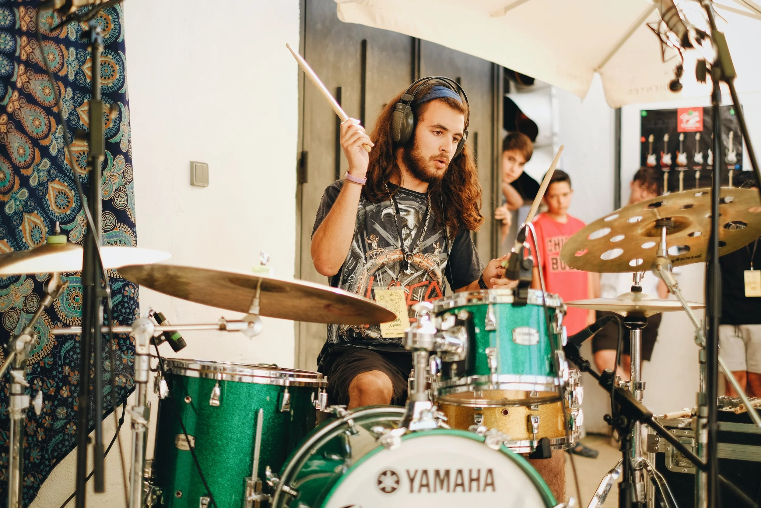 Joven tocando la batería en un estudio, con varias personas observando a su alrededor.