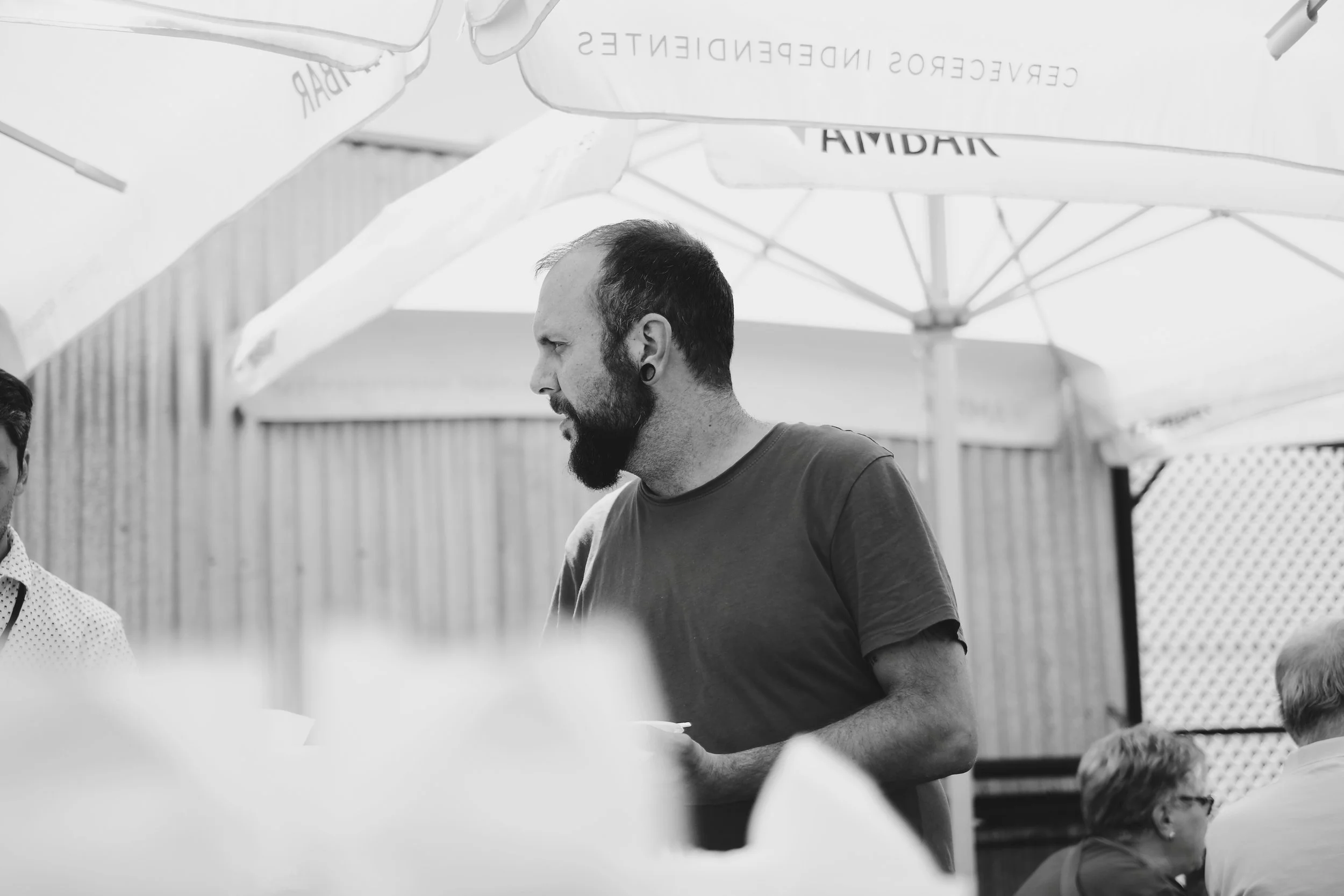 Hombre con barba y piercing en la oreja en un entorno al aire libre con paraguas y otras personas.