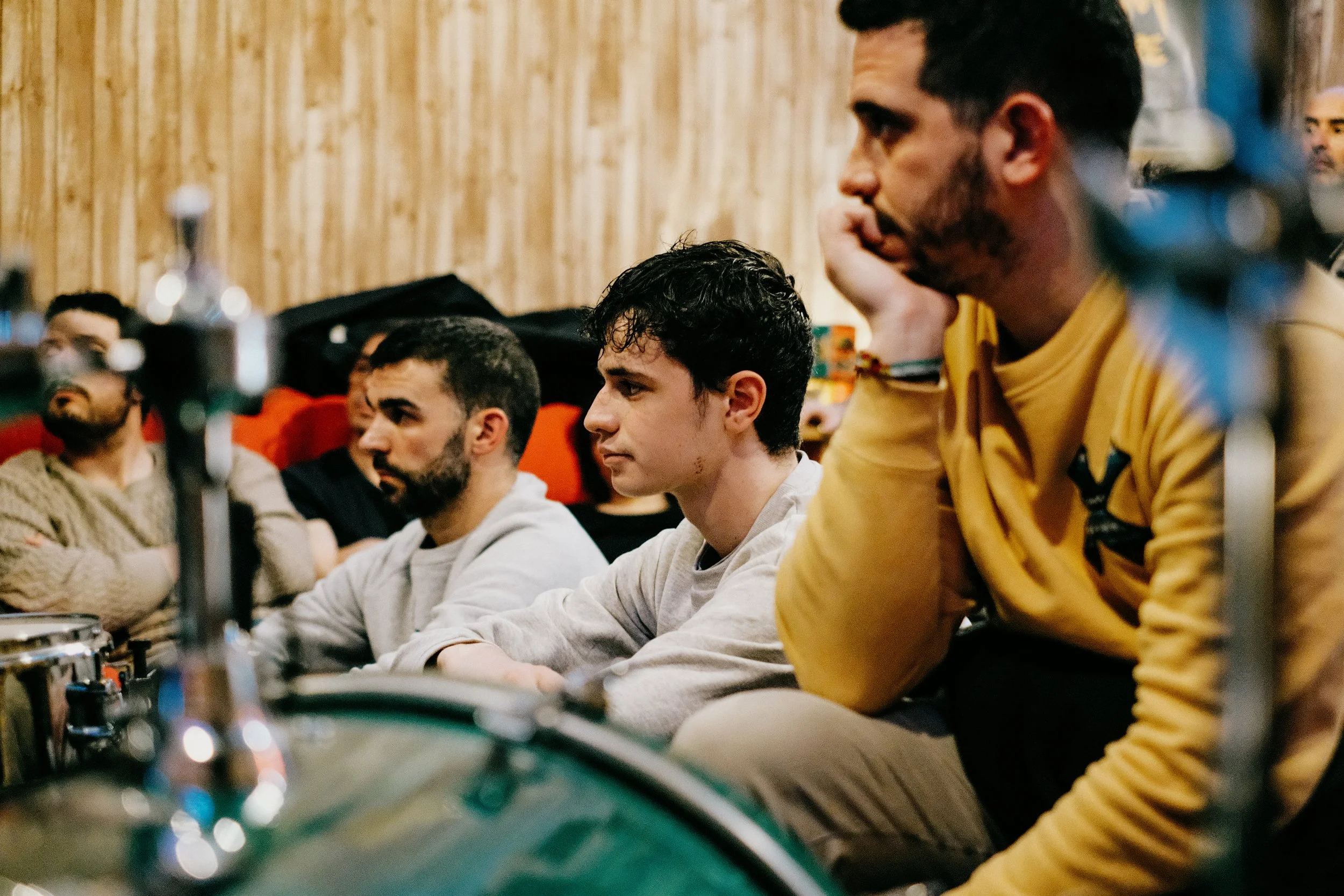 Grupo de personas sentado en una sala, escuchando atentamente, algunos con expresión pensativa, en un ambiente interior con paredes de madera.