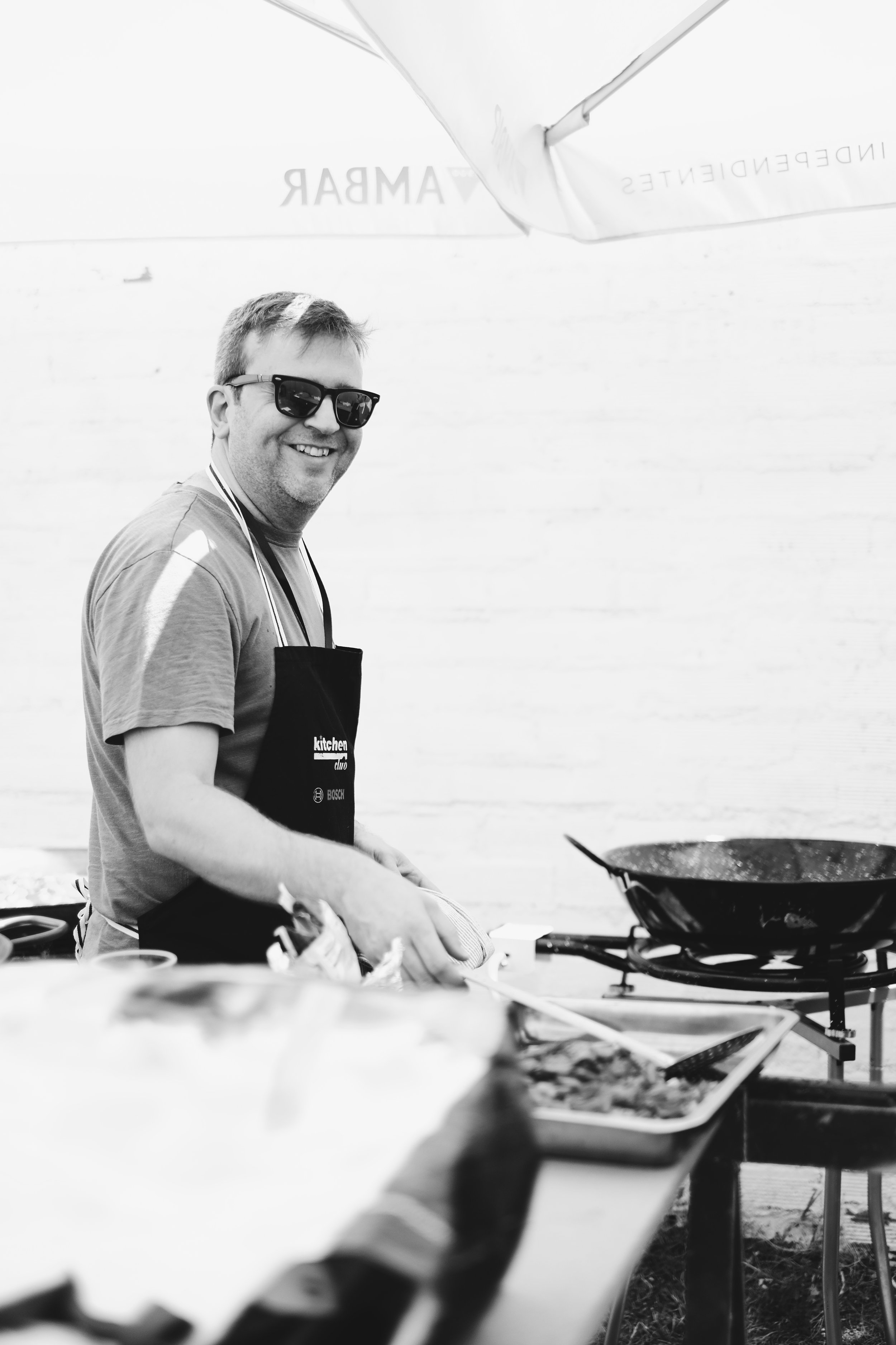 Hombre sonriendo con gafas de sol, usando delantal, cocinando en una sartén al aire libre.