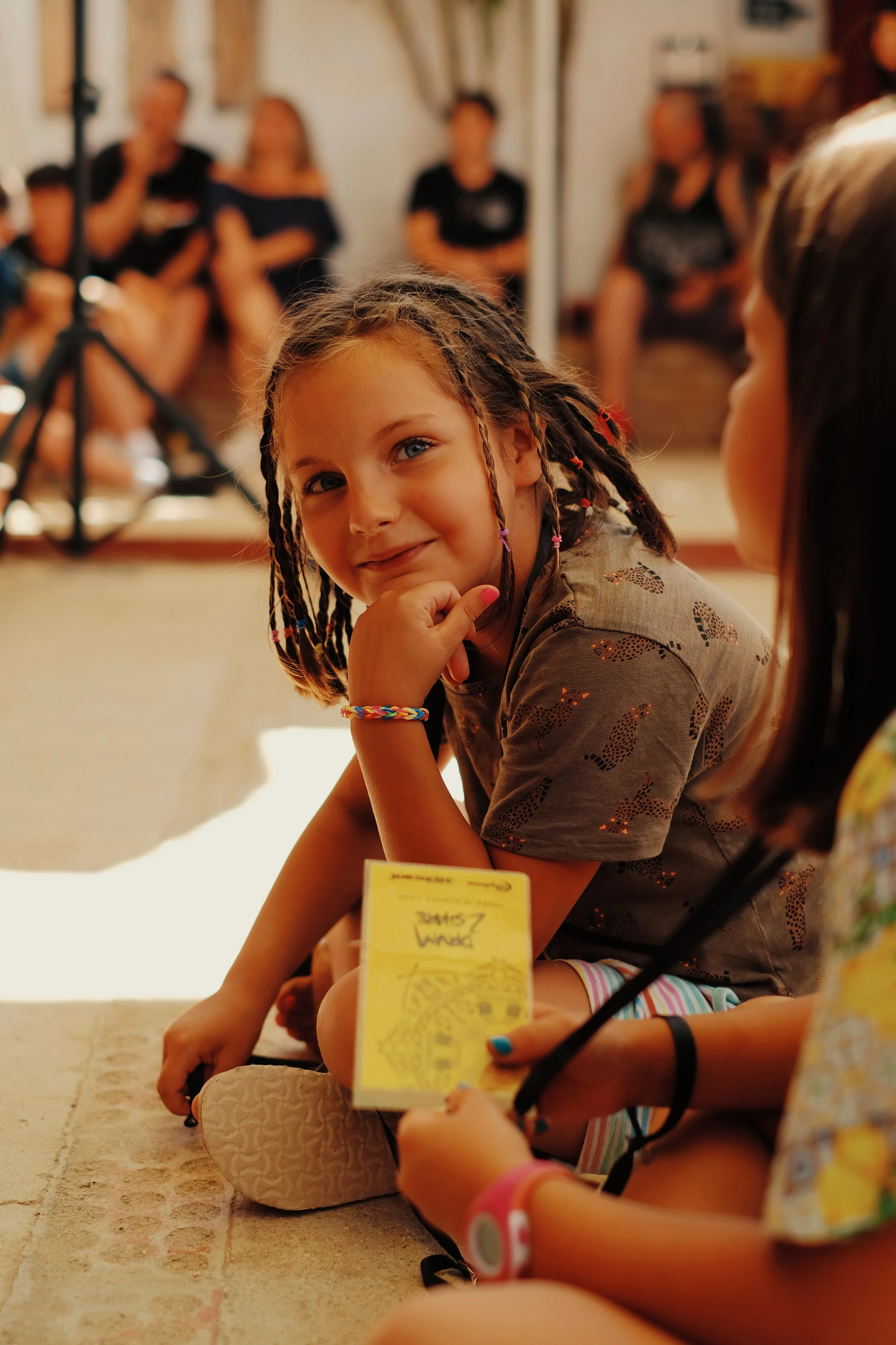 Niña de cabello trenzado con cara pensativa, sentada en el suelo en un ambiente interior, con otras personas en el fondo.