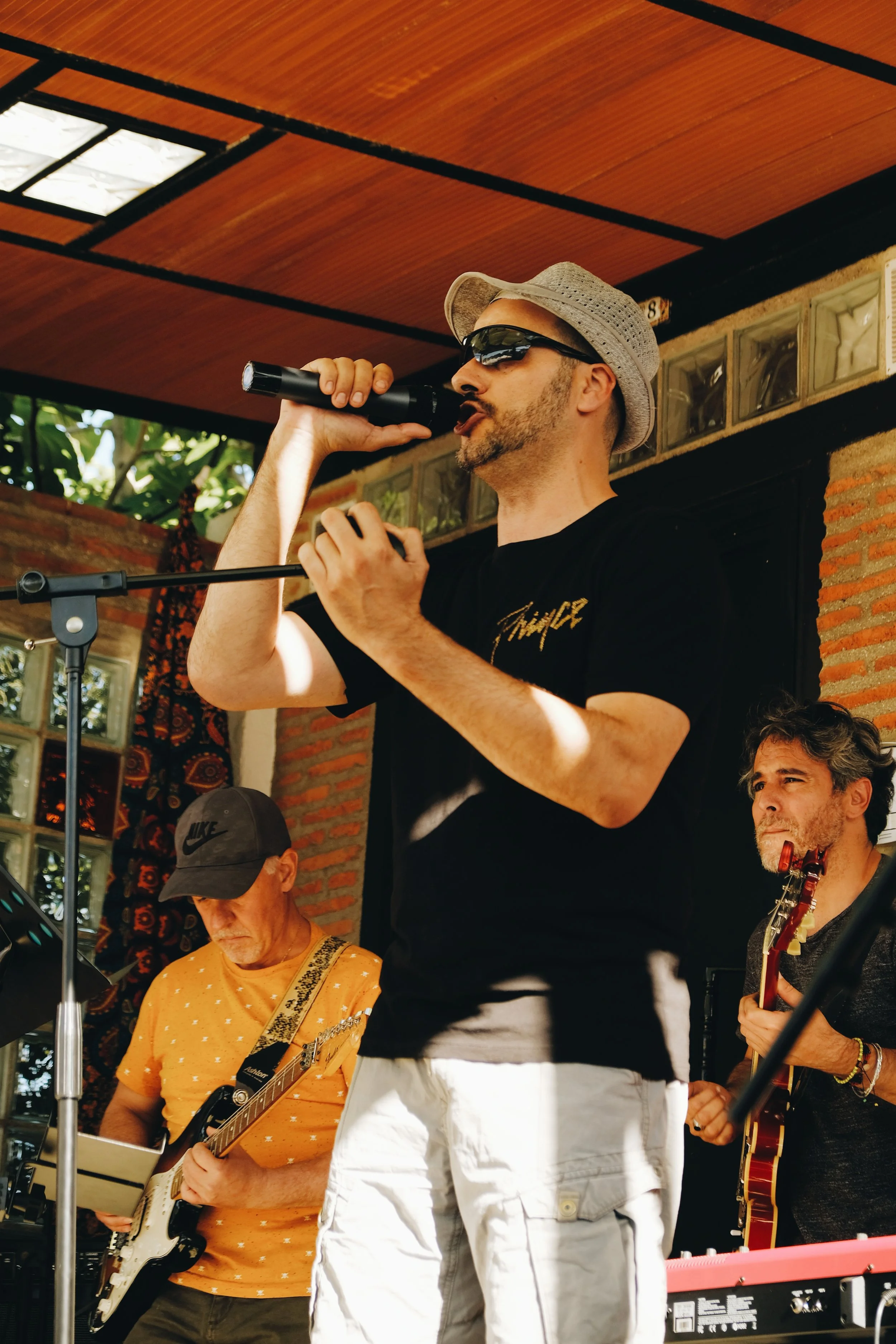 Grupo de músicos tocando en una presentación al aire libre, con un hombre en el centro cantando y tocando la armónica, acompañados de otros músicos con guitarra y teclado.