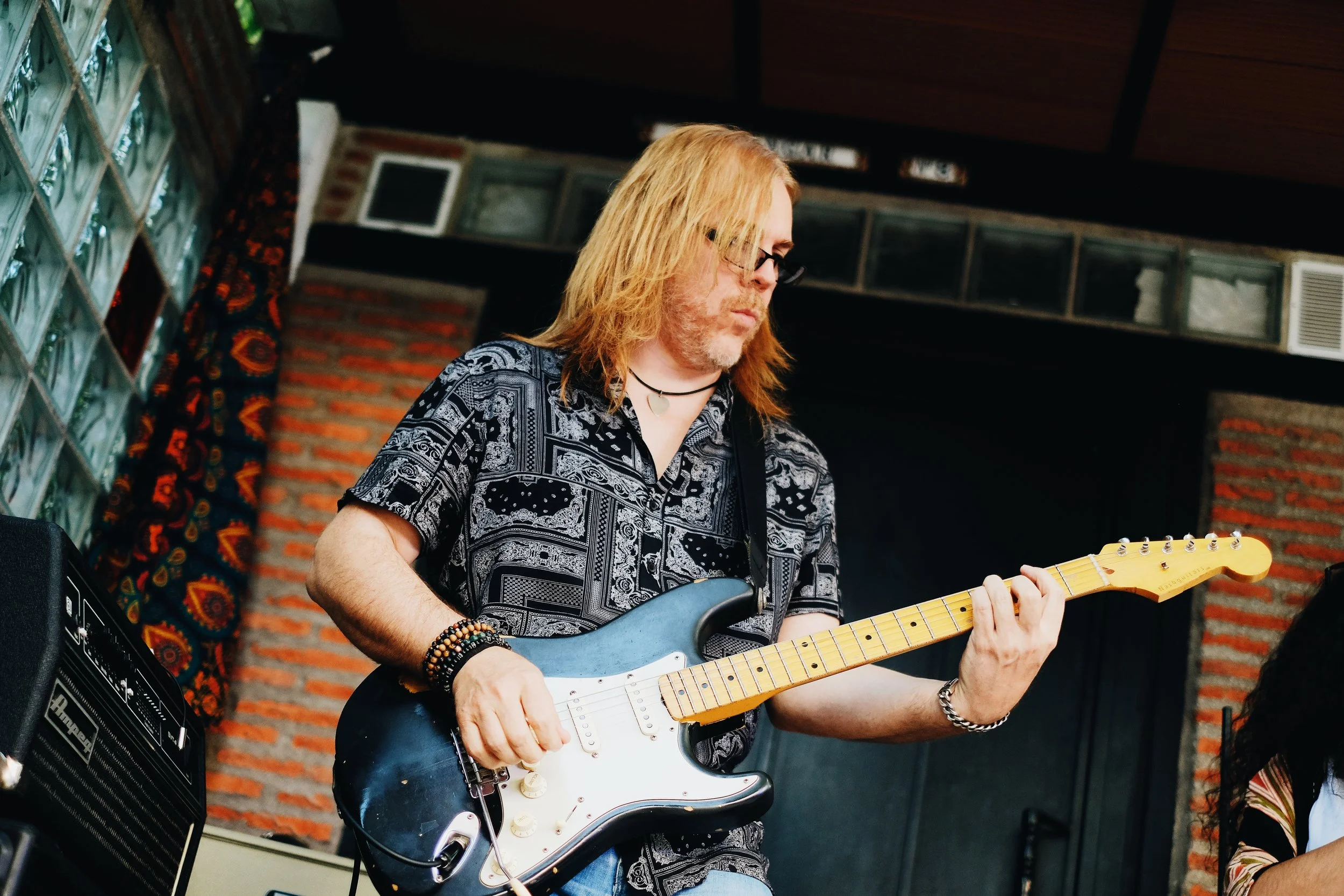 Un hombre con cabello largo rubio, gafas y una camiseta negra con patrones, toca una guitarra eléctrica en un escenario con paredes de ladrillo y decoraciones vintage.