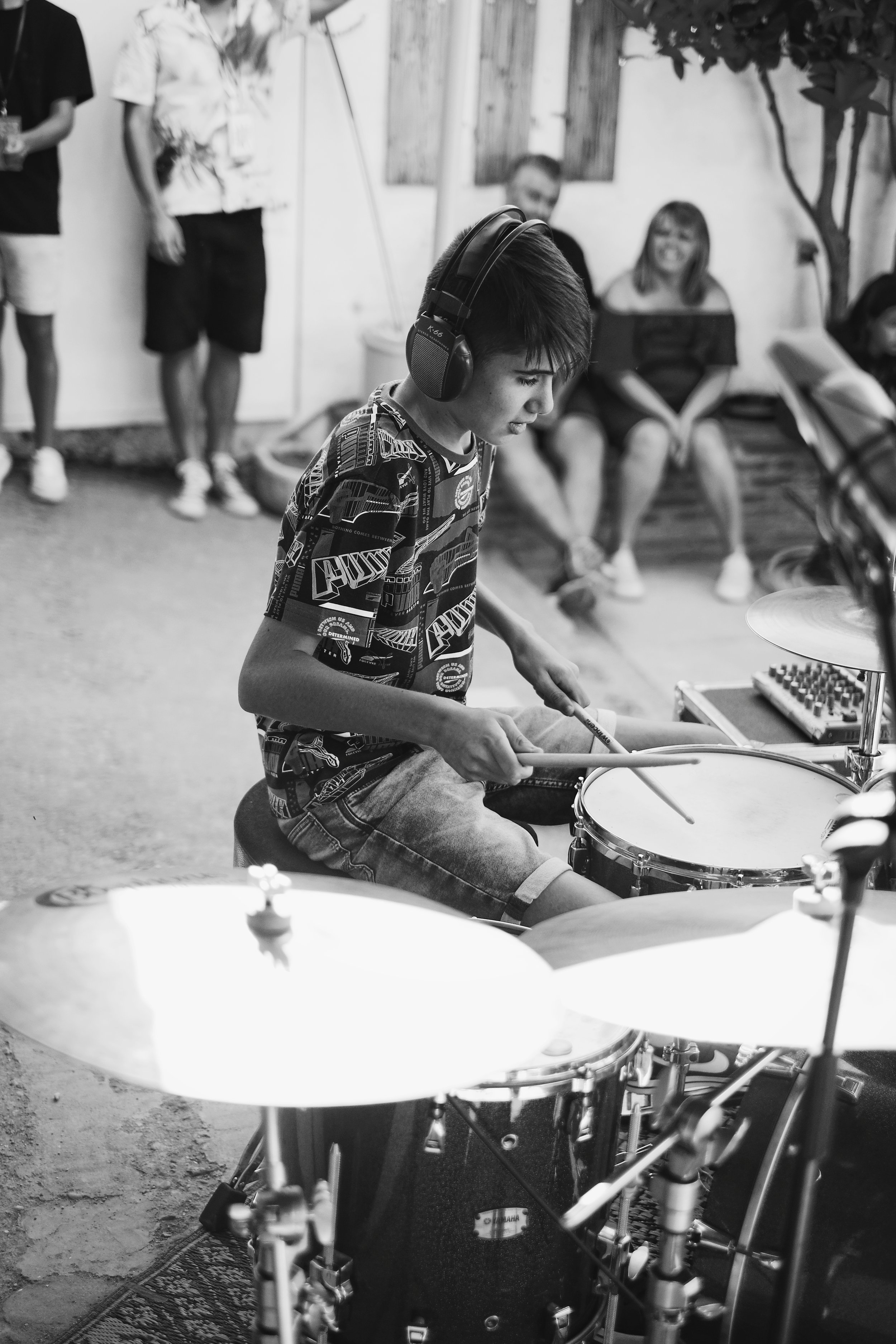 Un niño tocando la batería en un ensayo o concierto, lleva audífonos y una camiseta con diseños, mientras un grupo de personas observa en el fondo.