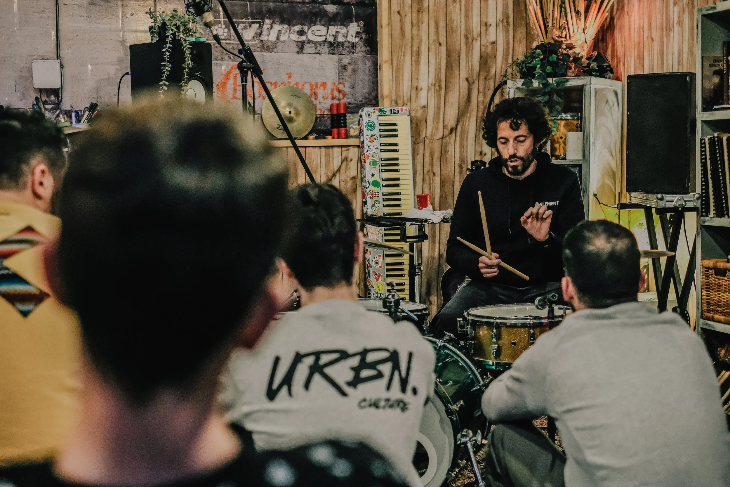 Un instructor de batería enseñando a un grupo de personas en un taller o clase en un ambiente interior con decoraciones coloridas y música.