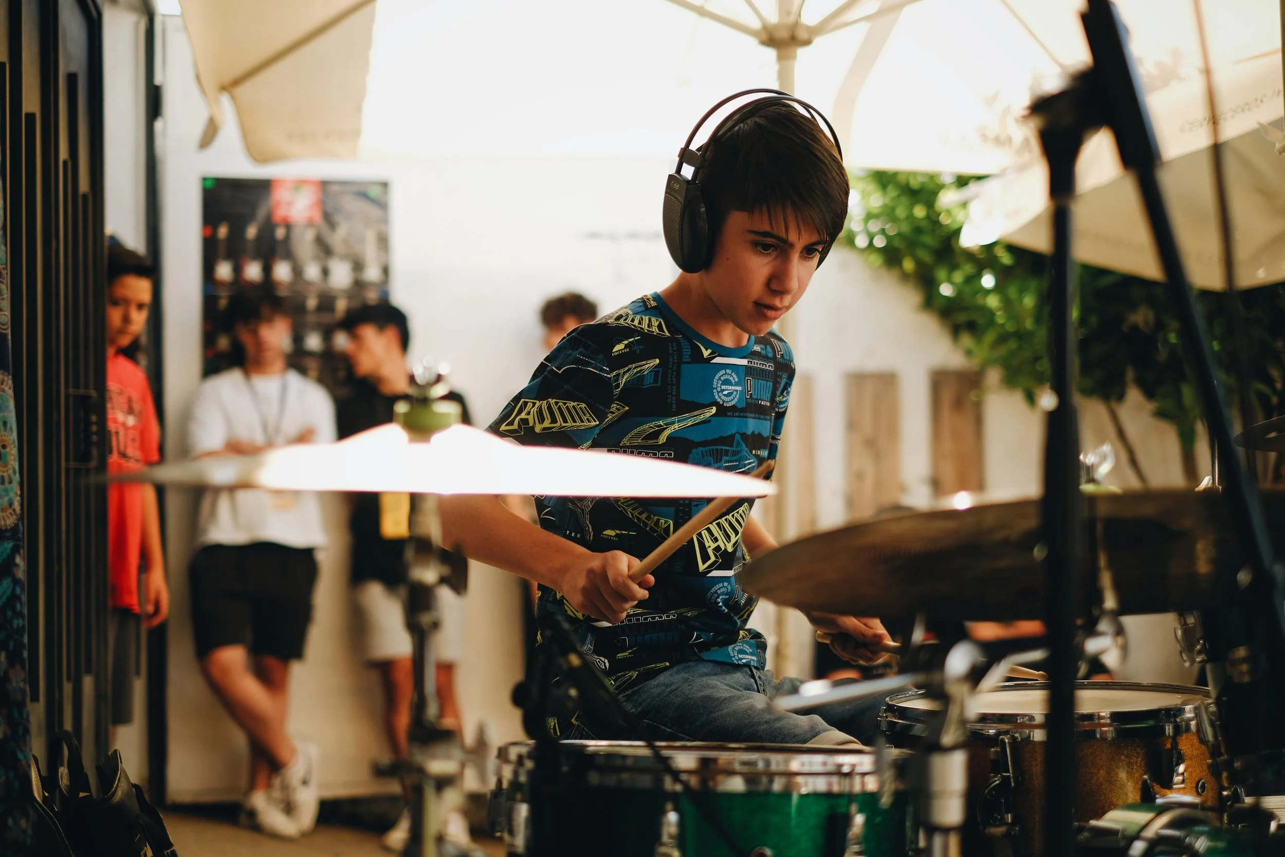 Joven tocando la batería con paraguas y bajo sombrillas, rodeado de personas observando.