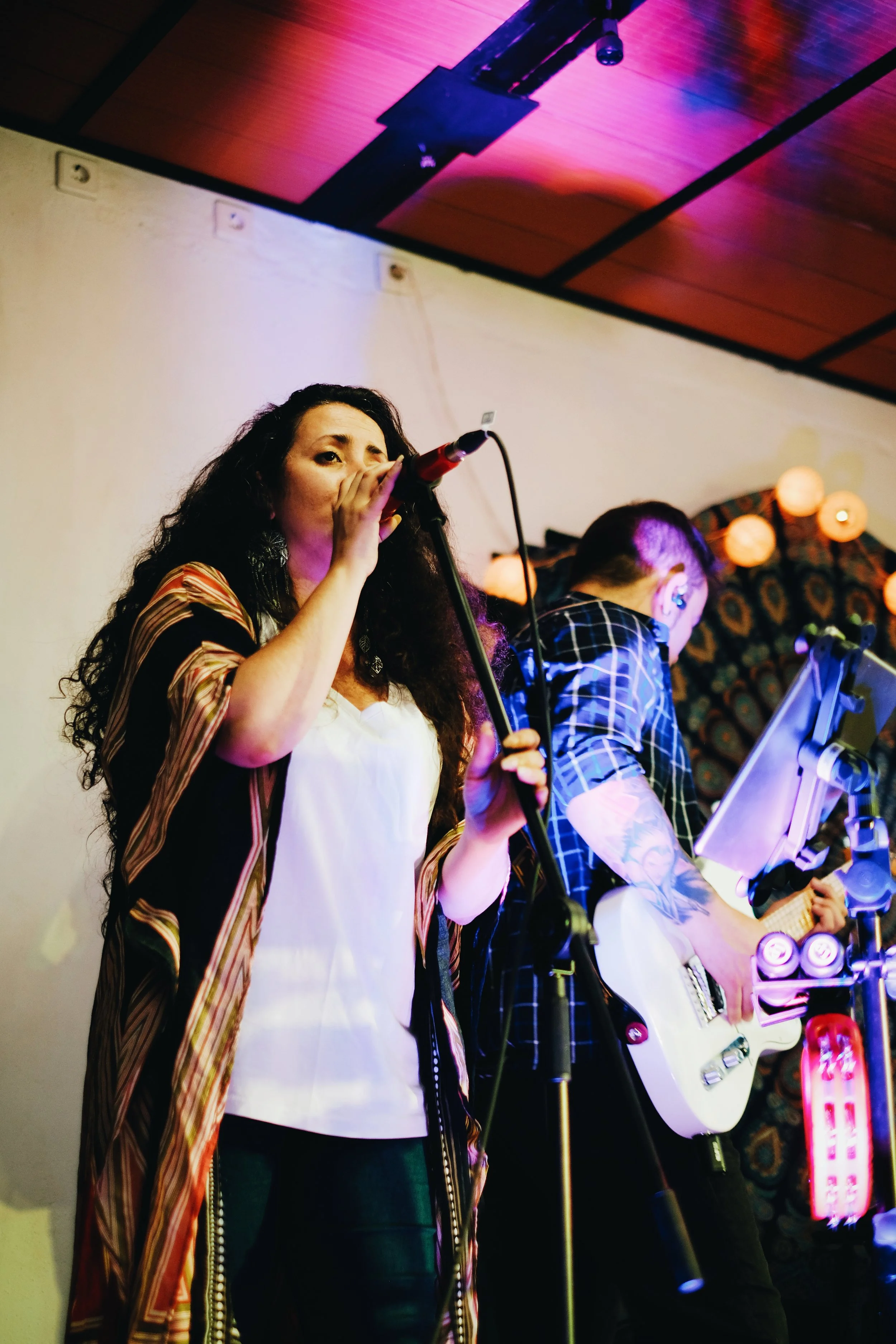 Mujer cantando con micrófono junto a un guitarrista en un escenario con iluminación de colores y decoraciones con luces de papel.