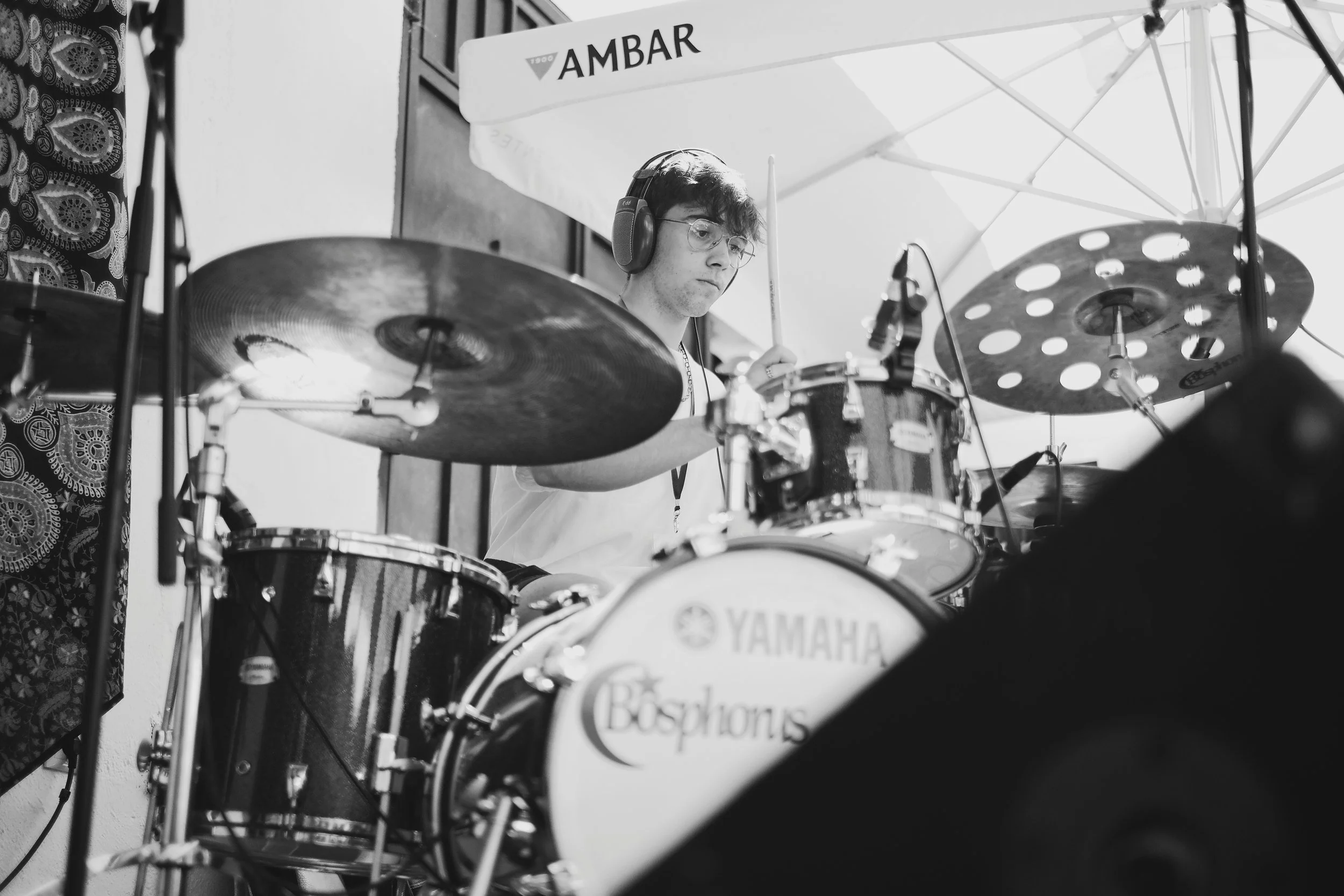 Joven tocando batería en un escenario con sombrilla y auriculares, en ambiente al aire libre en un día soleado.