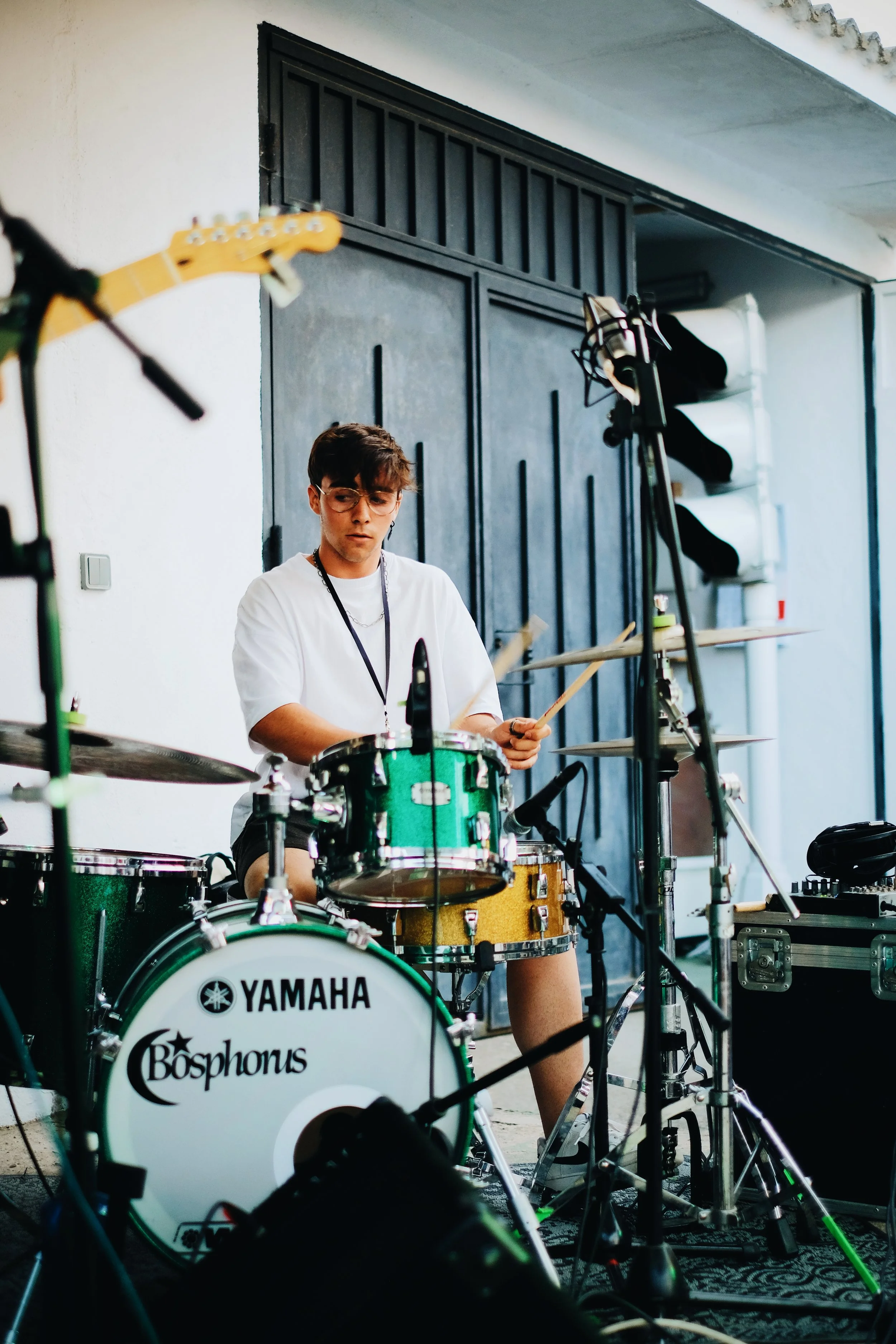 Joven tocando batería en un escenario, rodeado de equipo musical y micrófonos.