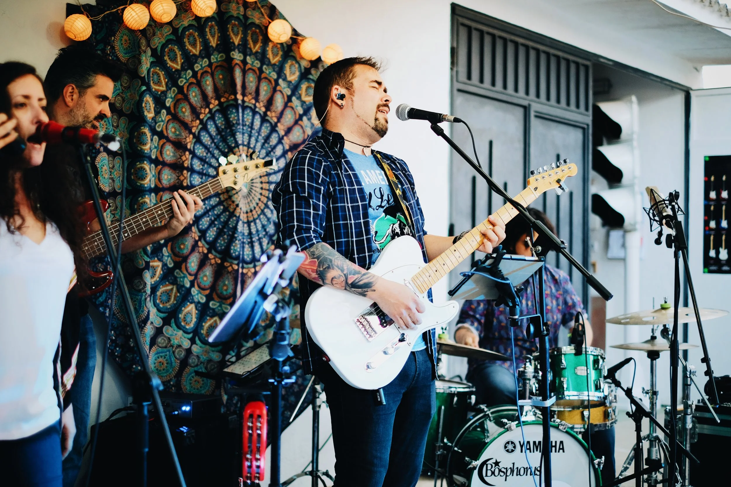 Un grupo de músicos tocando en un escenario, con instrumentos como guitarra eléctrica, bajo y batería, en un entorno decorado con luces de papel y un tapiz de fondo colorido.