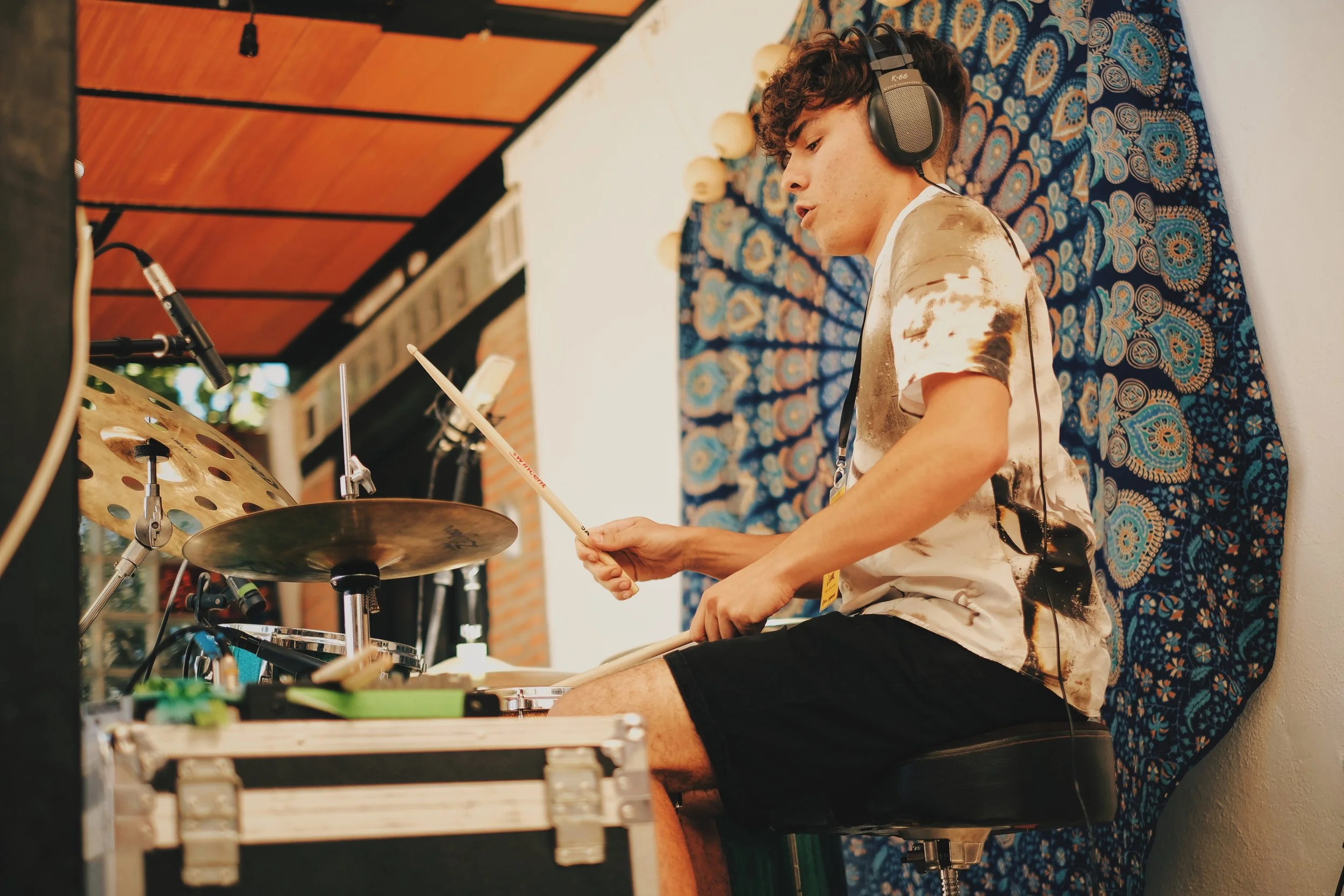 Joven tocando la batería mientras usa audífonos y una camiseta de manga corta con manchas. Está sentado en un taburete con telón de fondo de tela azul con patrones. Tiene un micrófono cerca y hay equipo de sonido y una caja de madera en primer plano.