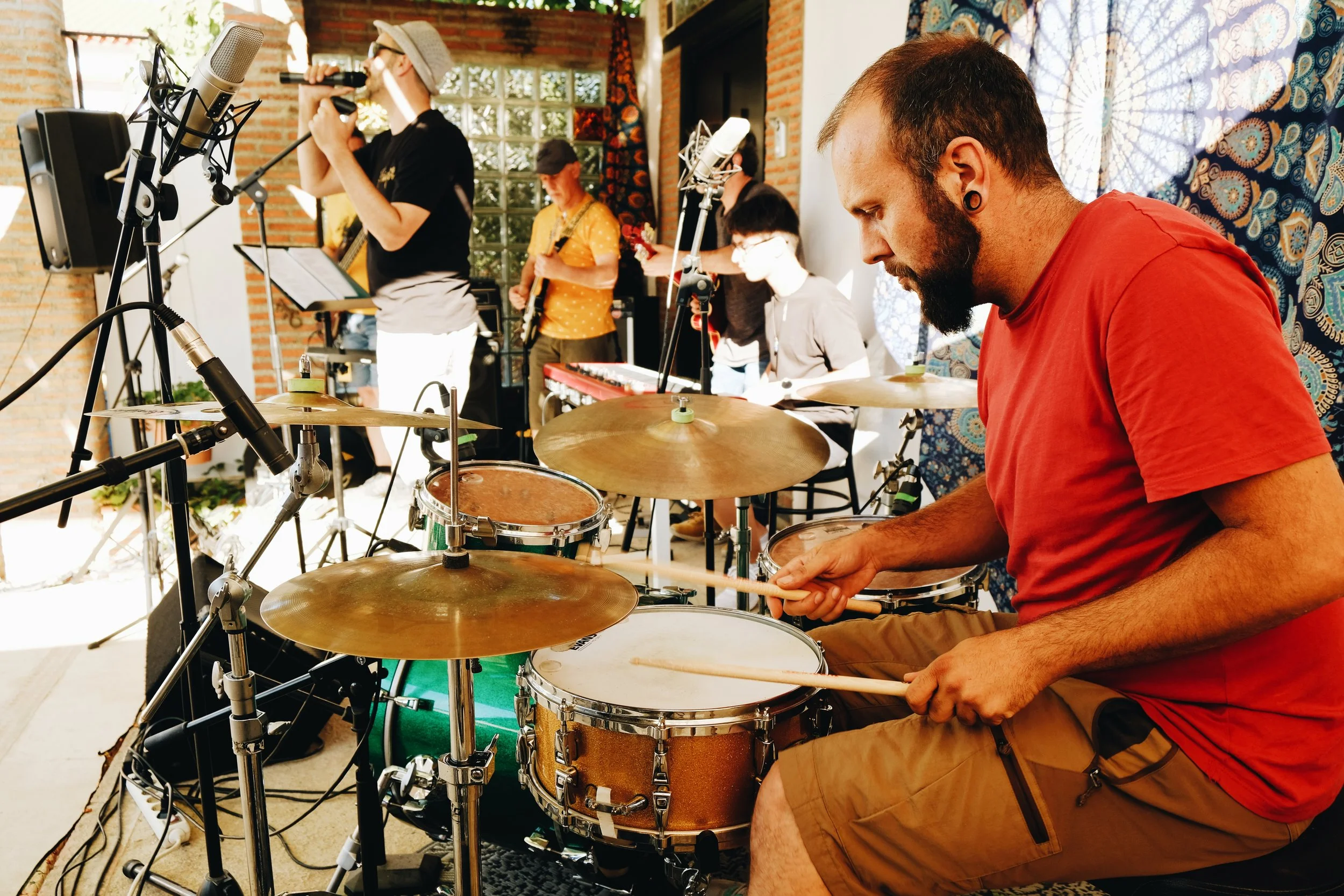 Un grupo de músicos tocando en un estudio, con un baterista en primer plano, otros músicos tocando guitarra y otros instrumentos en el fondo.