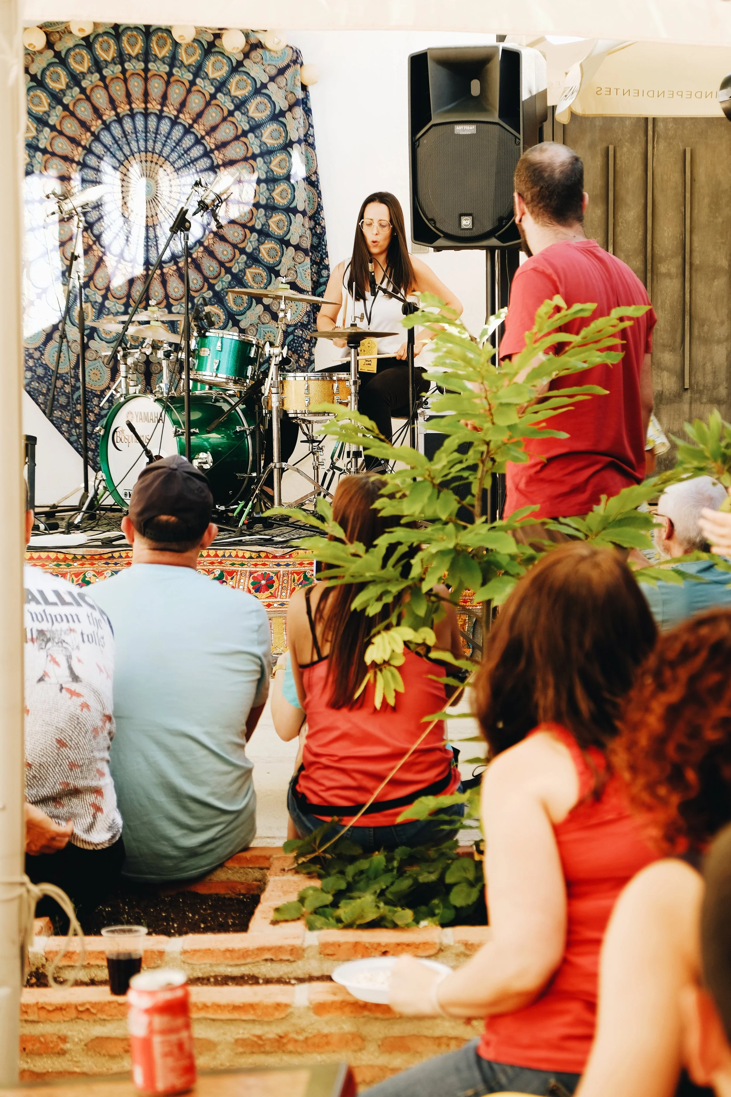 Una mujer tocando la batería en una presentación al aire libre, con un público viéndola mientras algunos están sentados en suelo y otros de pie, en un ambiente de concierto o evento cultural.