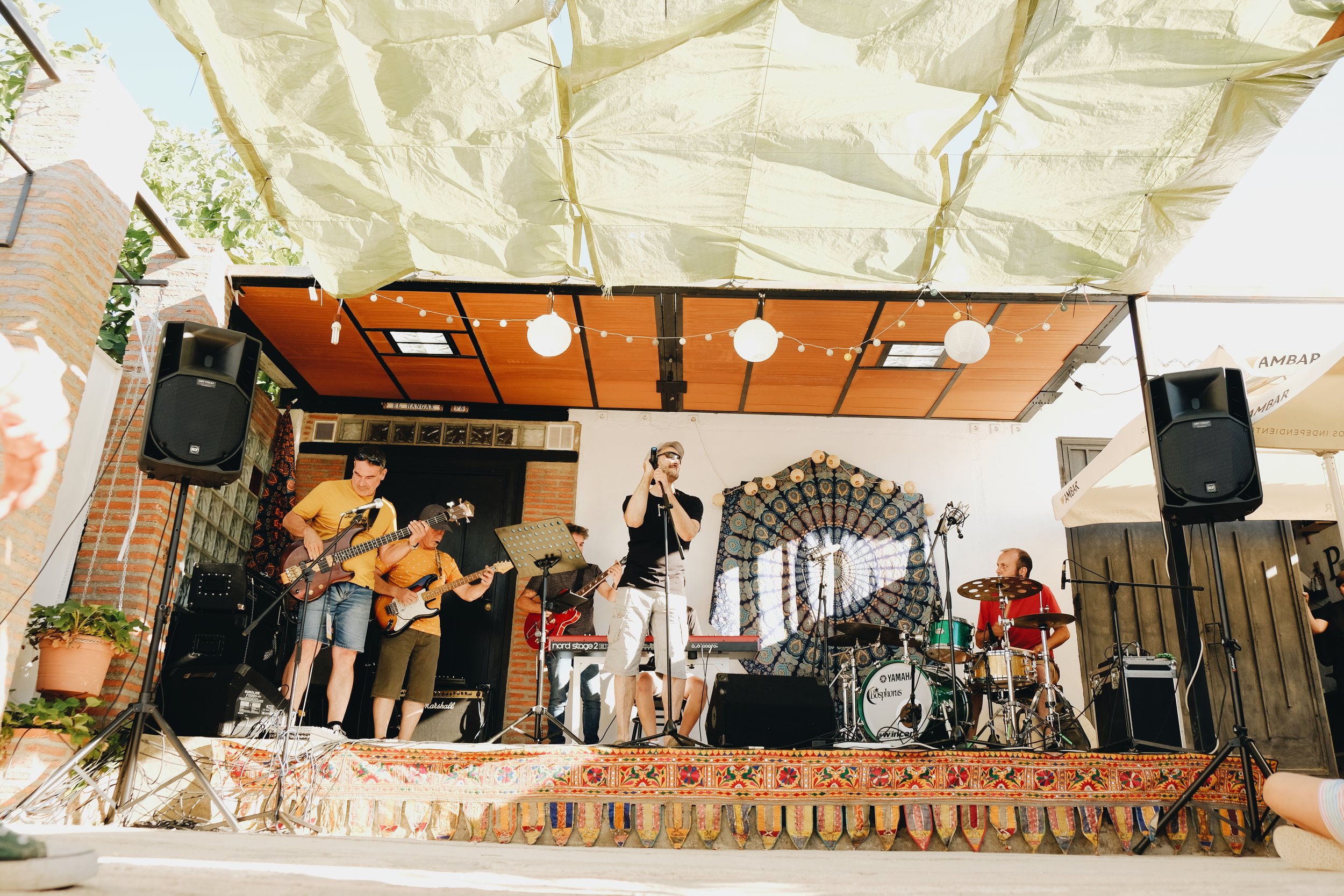 Una banda musical tocando en un escenario al aire libre, con varios músicos que cantan, tocan guitarras y batería, decoraciones bohemias en el fondo y luces decorativas colgando del techo.