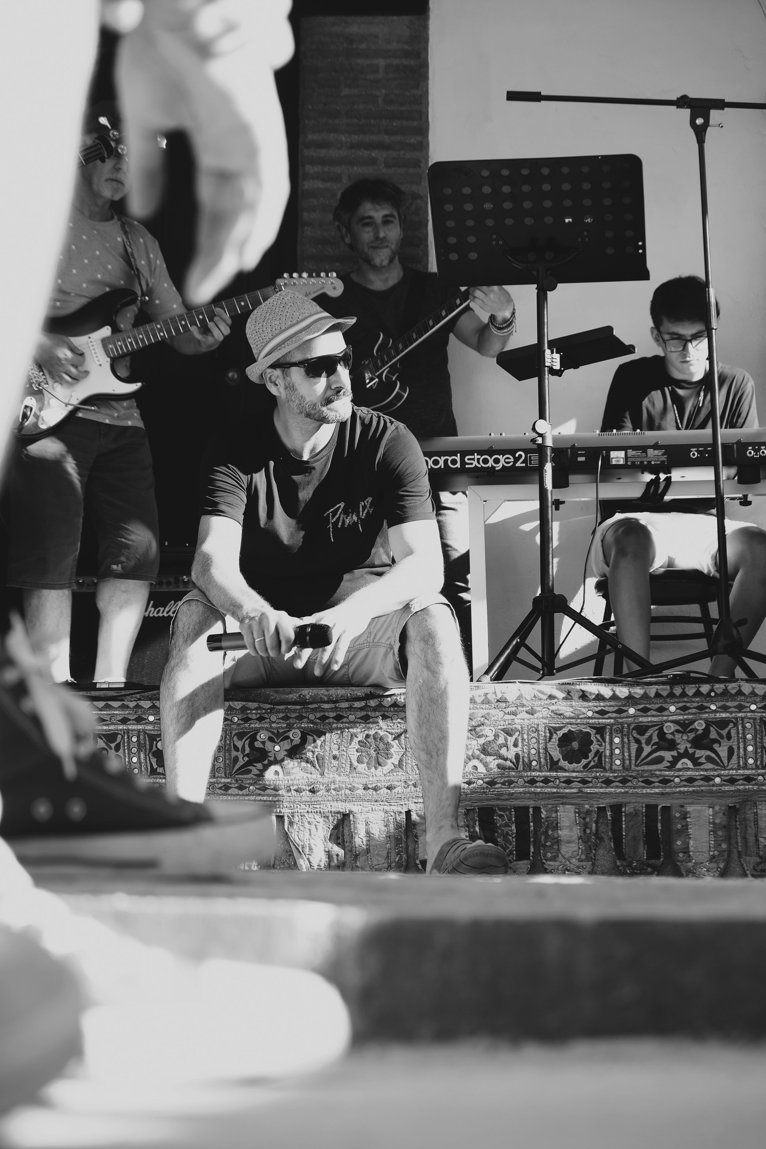 Grupo de músicos en un escenario, con uno sentado en el frente sosteniendo un micrófono y otros de pie tocando guitarras y teclado, en un ambiente de concierto.