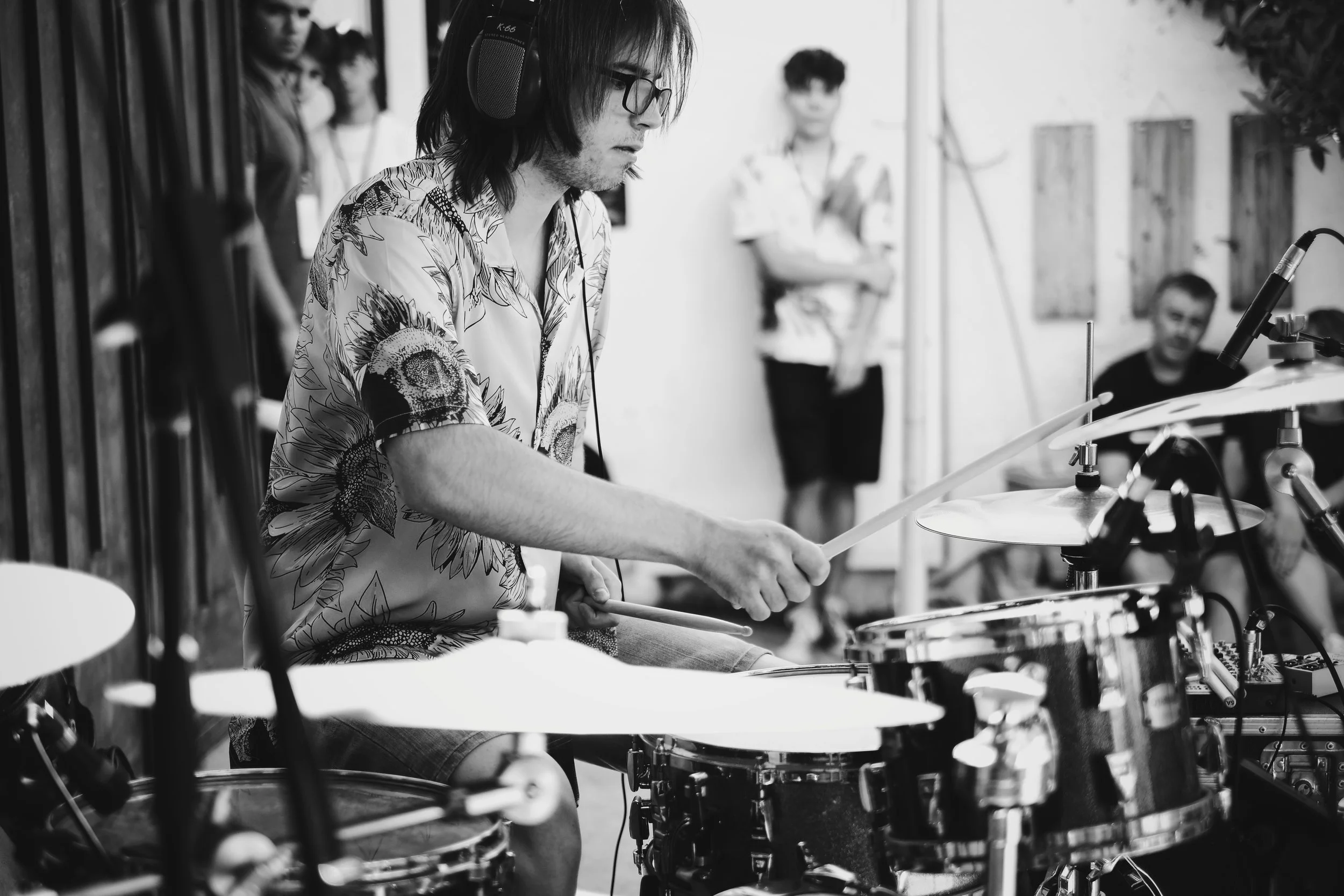 Hombre tocando la batería en un estudio o escenario, con personas observando al fondo.