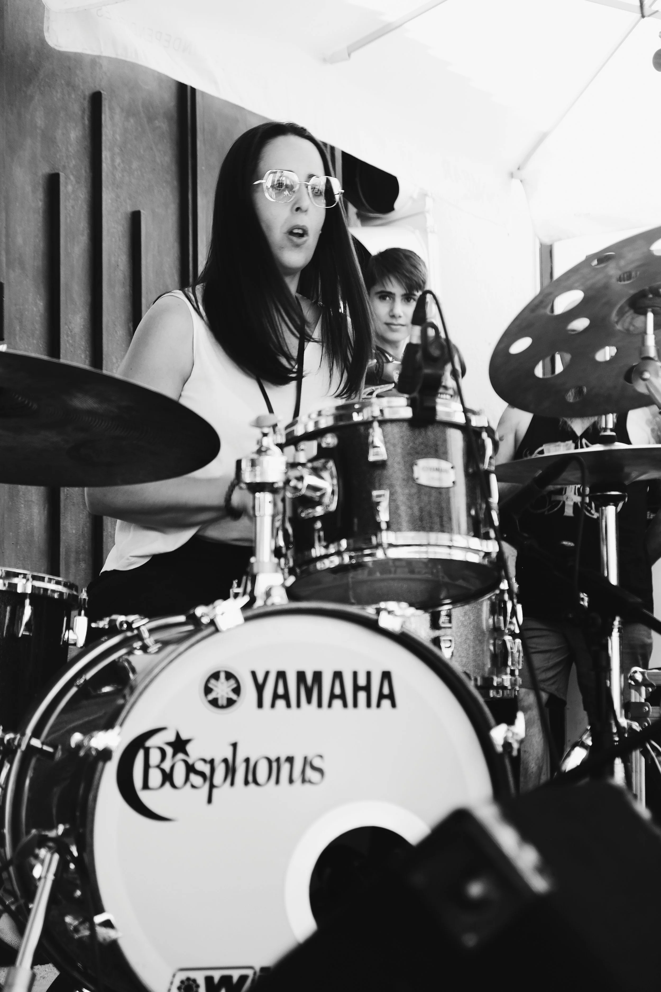 Mujer tocando la batería durante una presentación. La batería tiene el logo 'Yamaha' y 'Bosphorus'. En el fondo, hay un joven observando. La imagen está en blanco y negro.