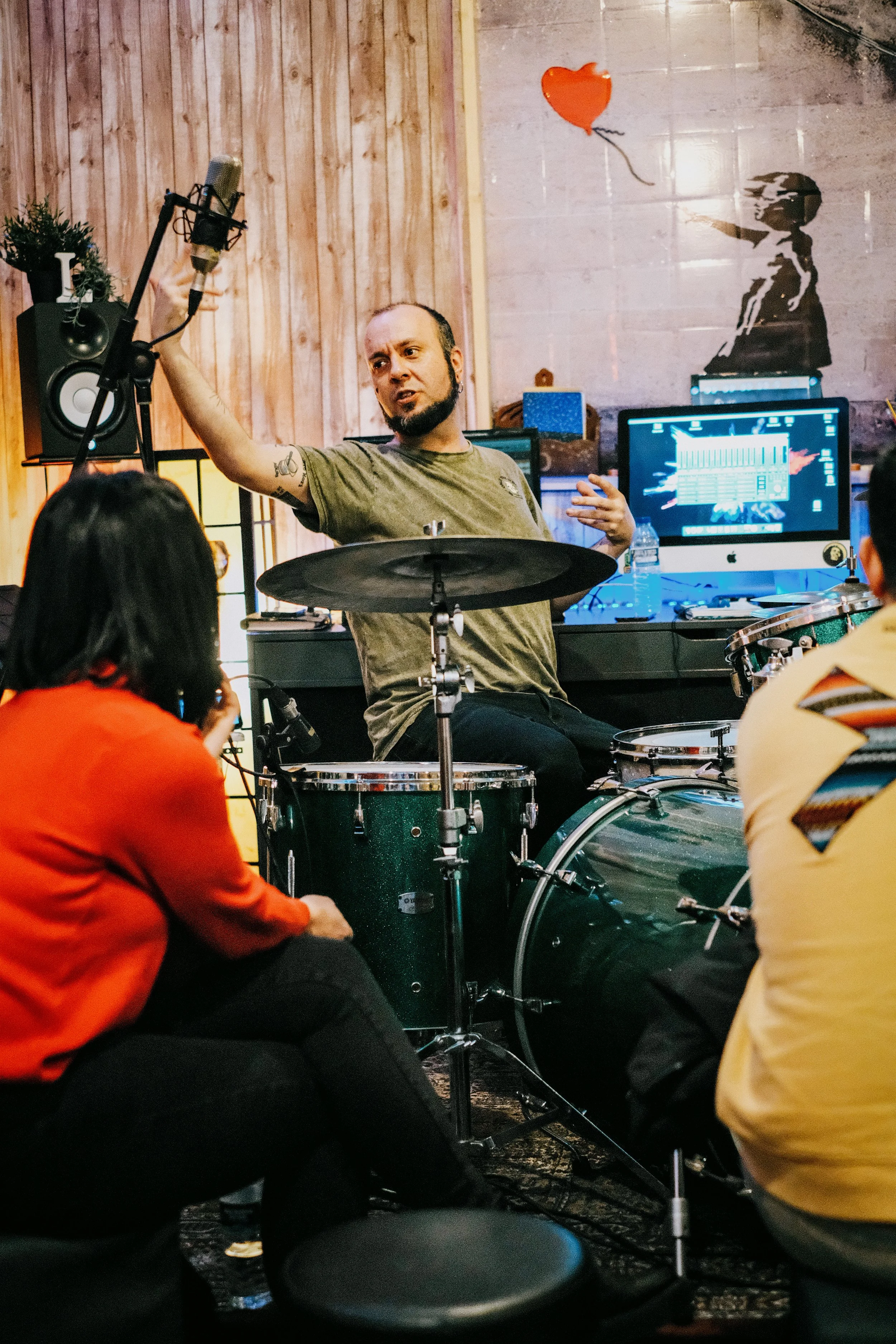 Un hombre tocando la batería en una sala de grabación rodeado de personas que lo observan.