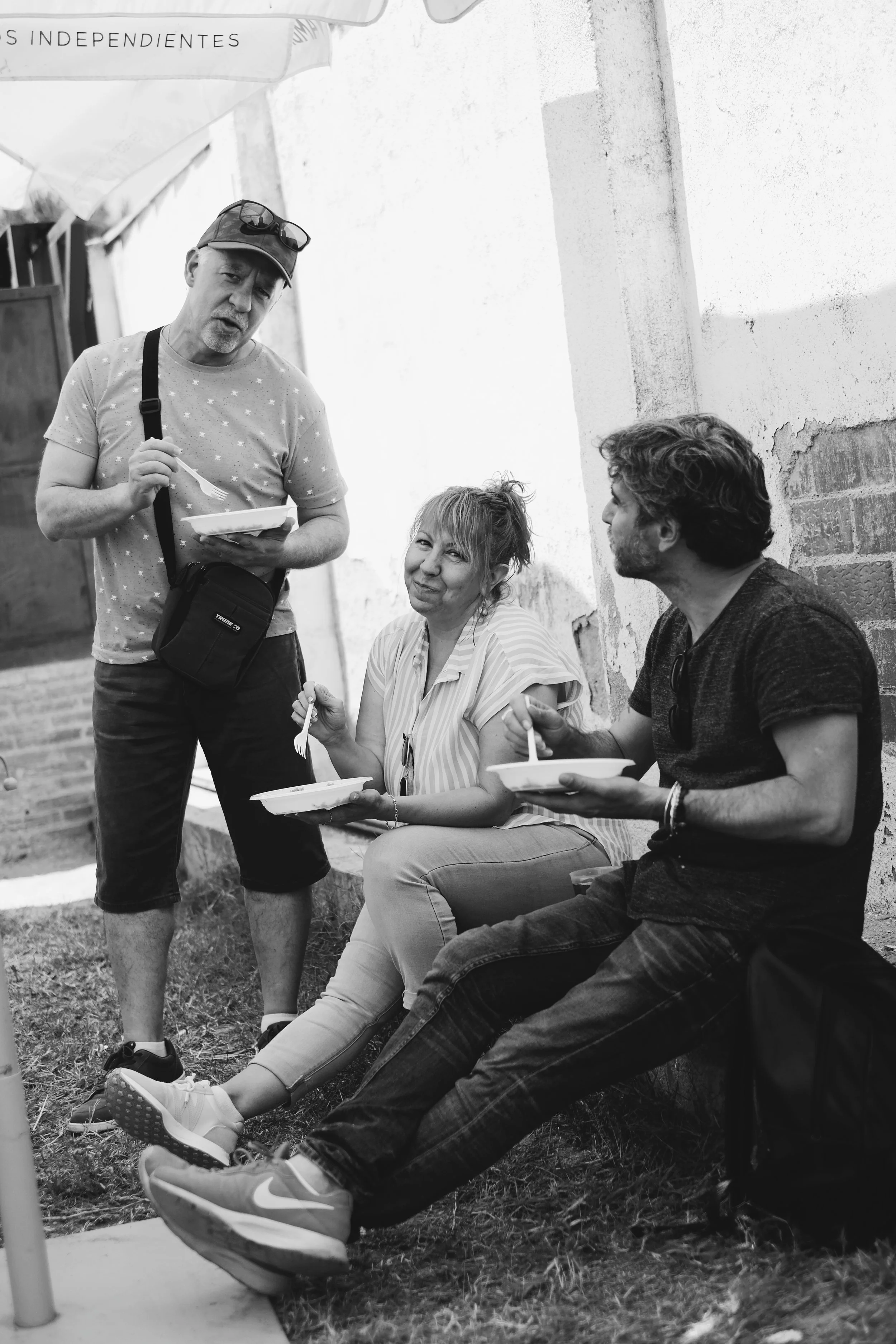Tres personas sentadas en una calle, dos con platos de comida y una con una bandeja, conversando y la mujer sonriendo.