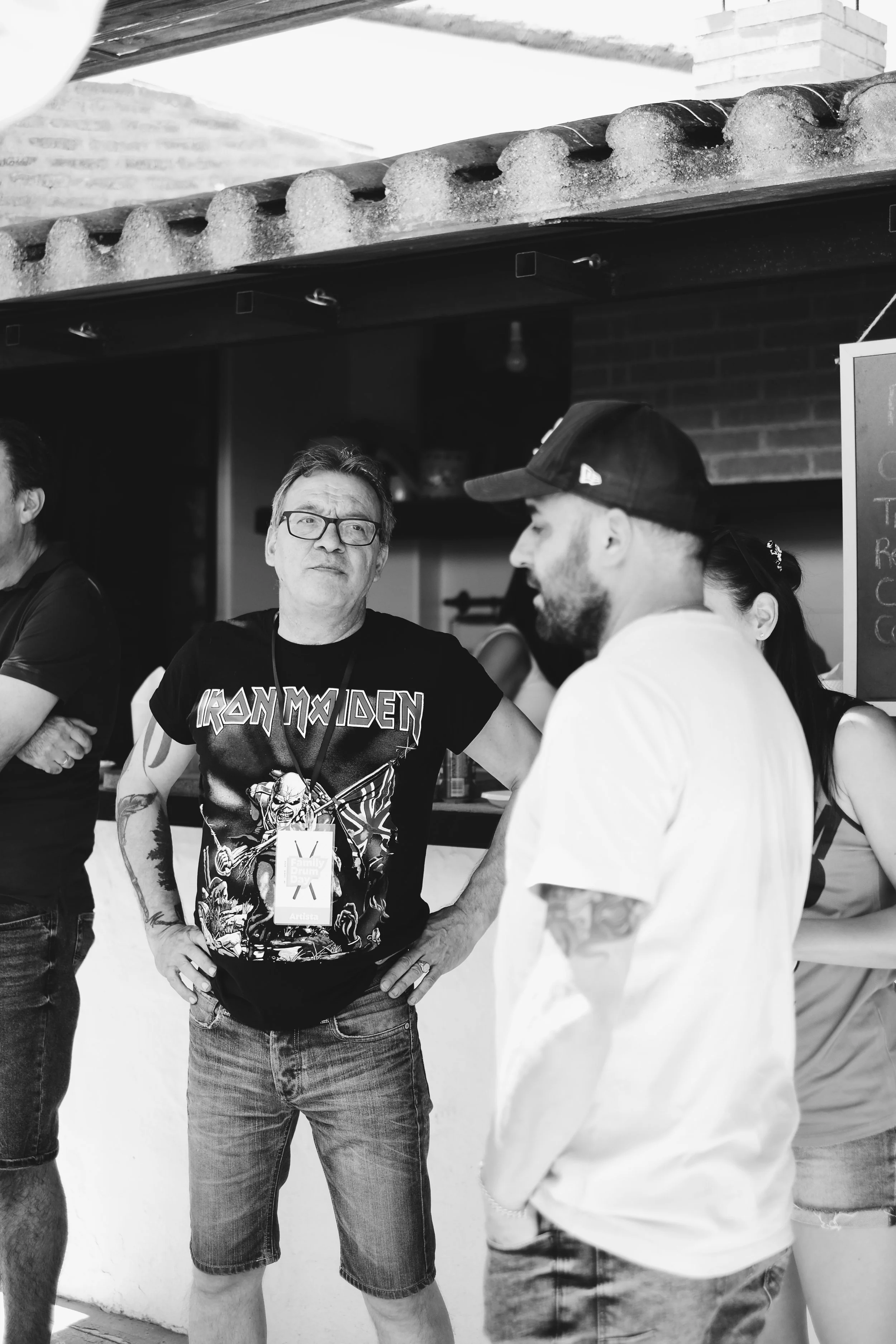 Personas conversando en un entorno informal, uno lleva una camiseta de Iron Maiden y usa gafas, mientras que otro lleva una gorra y tiene tatuajes en el brazo, en un espacio que parece ser un restaurante o cafetería.