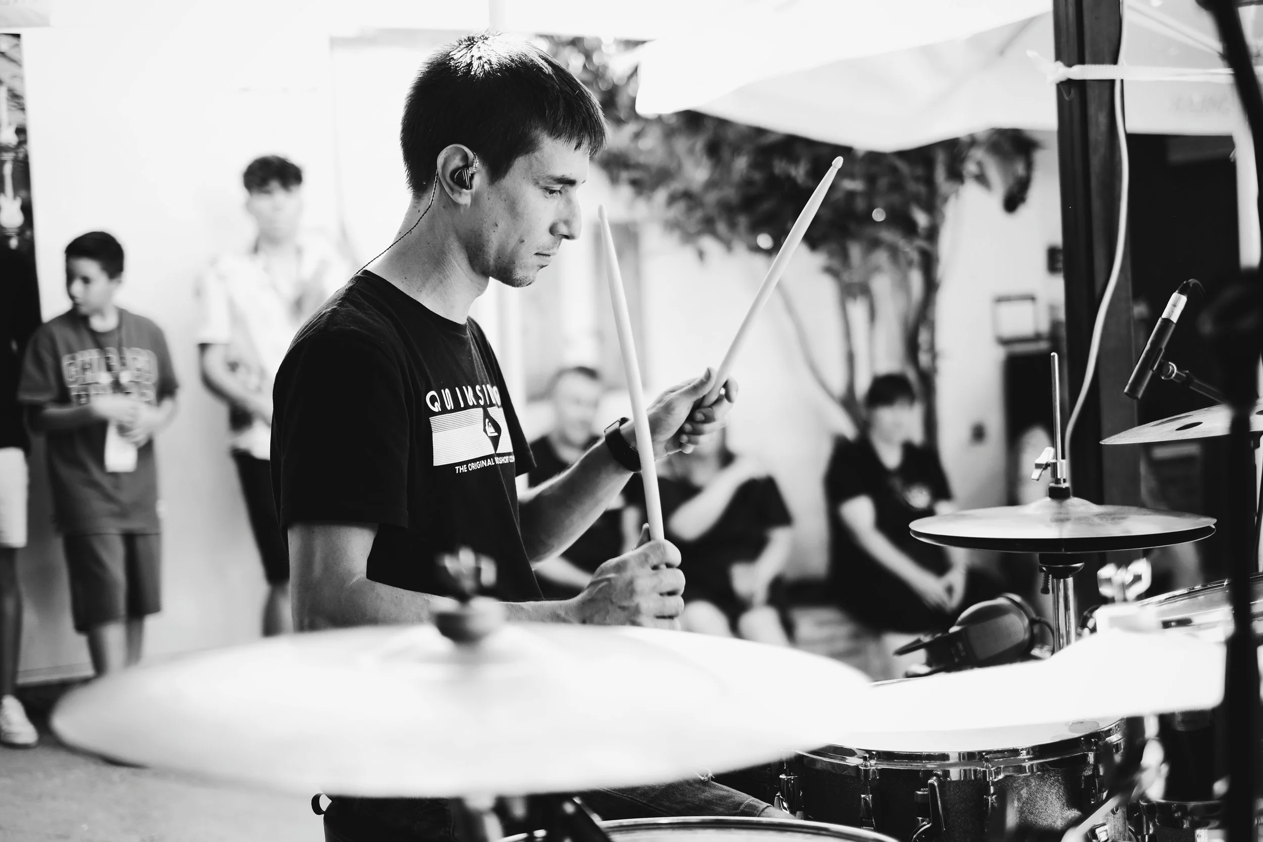 Hombre tocando la batería en un evento, con varias personas observando en el fondo, en blanco y negro.