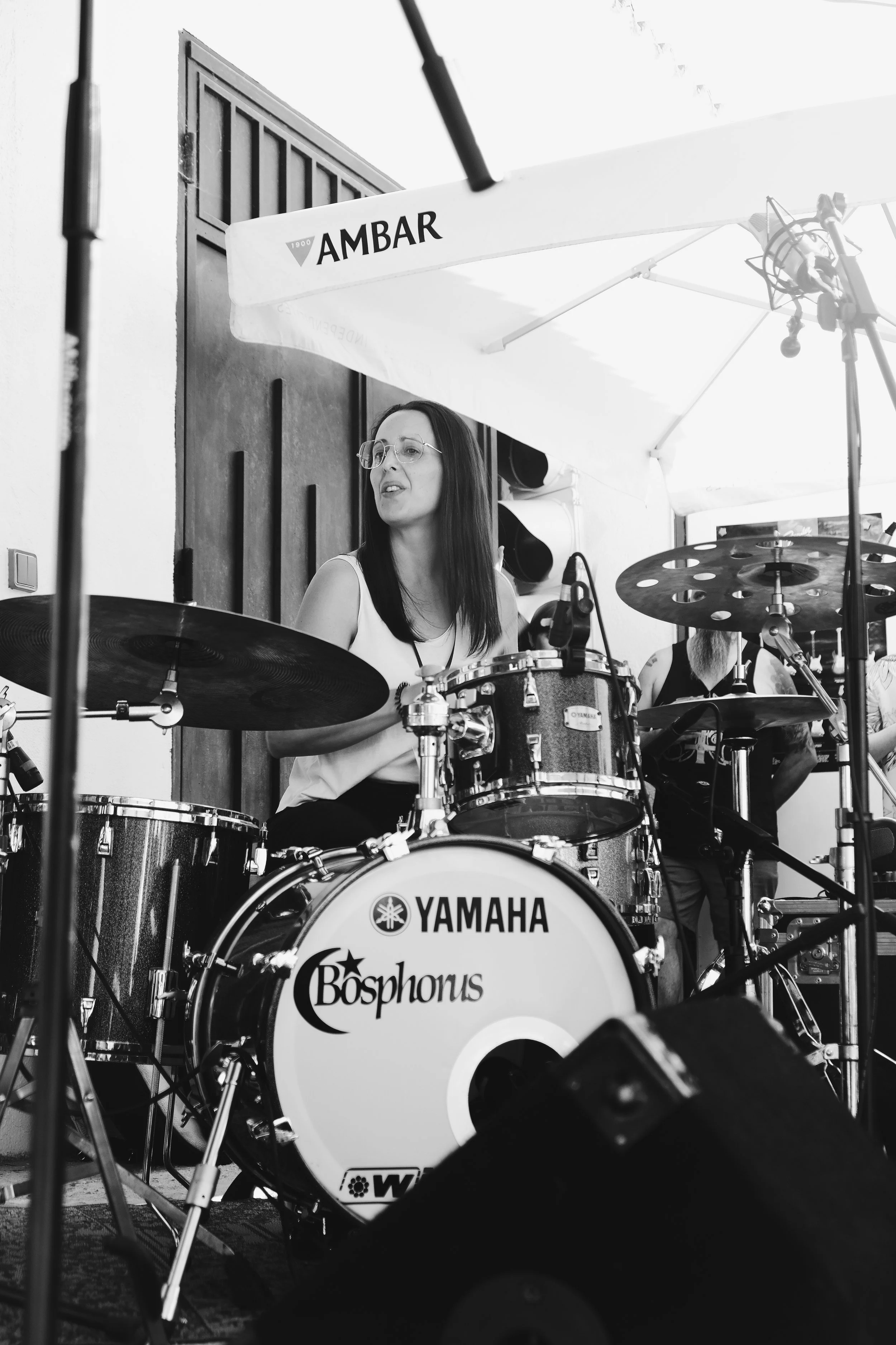 Mujer tocando la batería en un escenario, con paraguas que tiene la marca 'AMBAR'.
