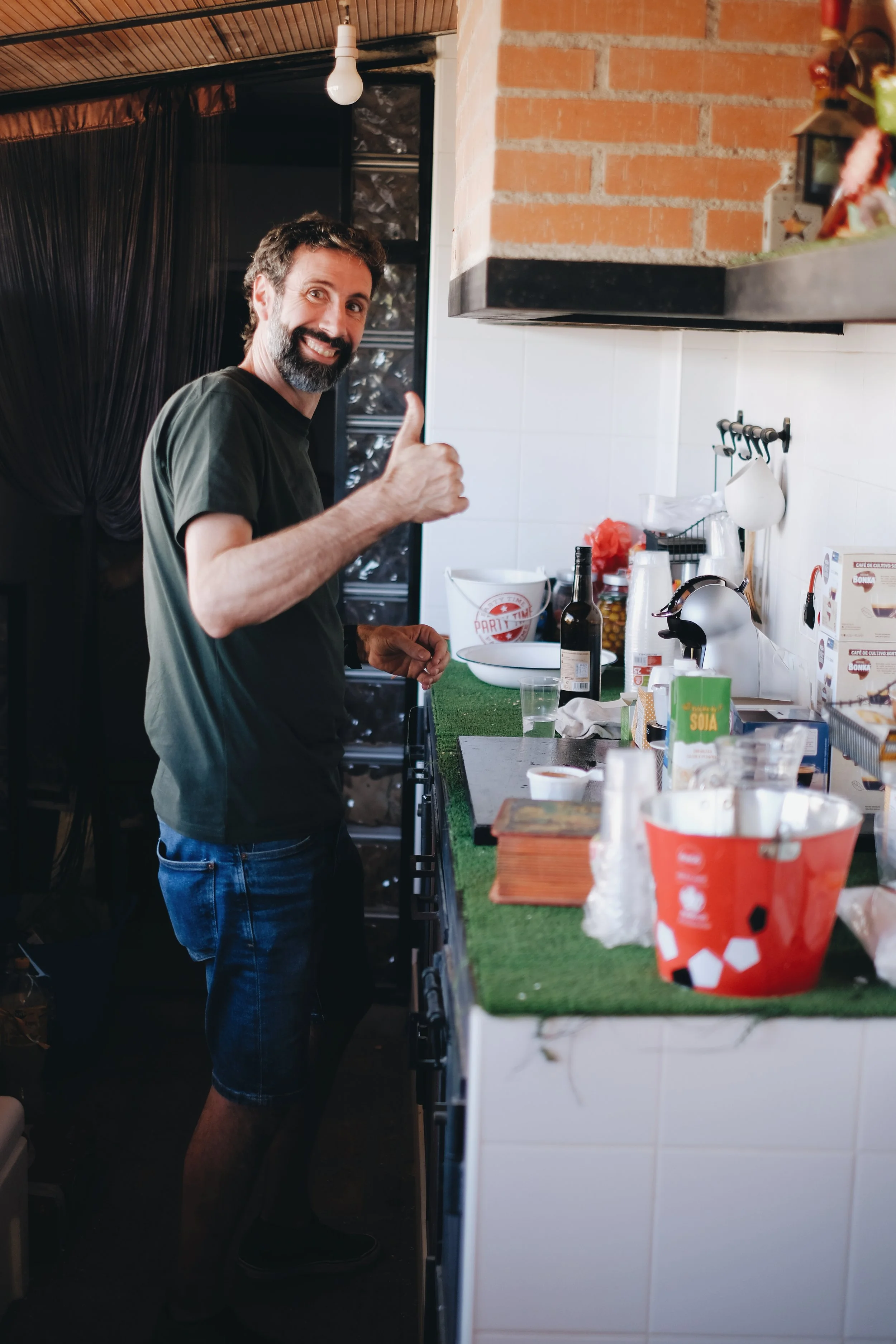 Hombre sonriendo y mostrando pulgar hacia arriba en una cocina. Hay botellas, platos y utensilios en la encimera.