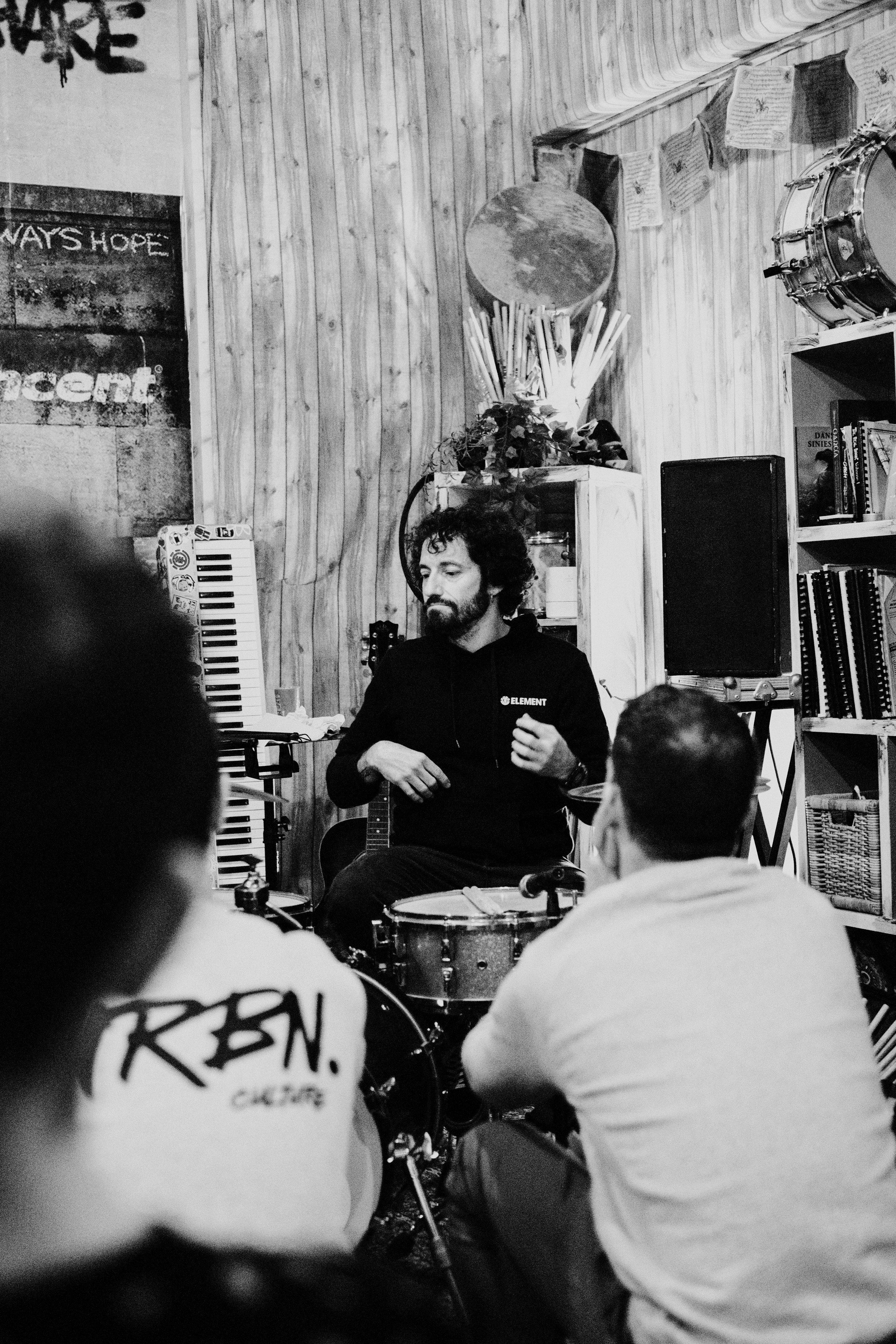 Un hombre con barba tocando la batería mientras un grupo de personas lo escucha en un ambiente íntimo de madera, decorado con instrumentos y libros.