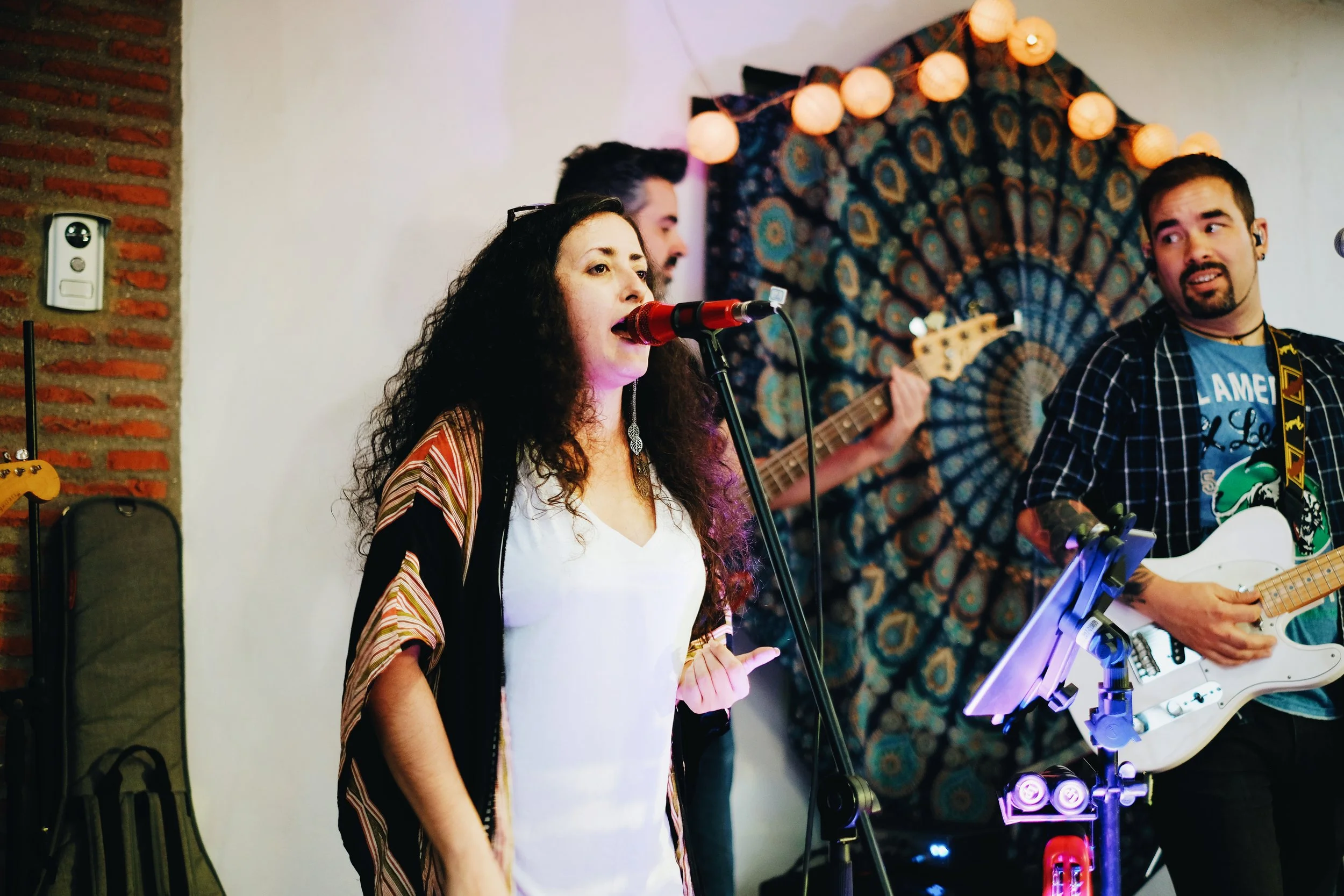 Grupo de músicos tocando en un escenario decorado con cuadros y luces. La mujer en el centro canta con un micrófono, mientras un hombre a la derecha toca la guitarra eléctrica.