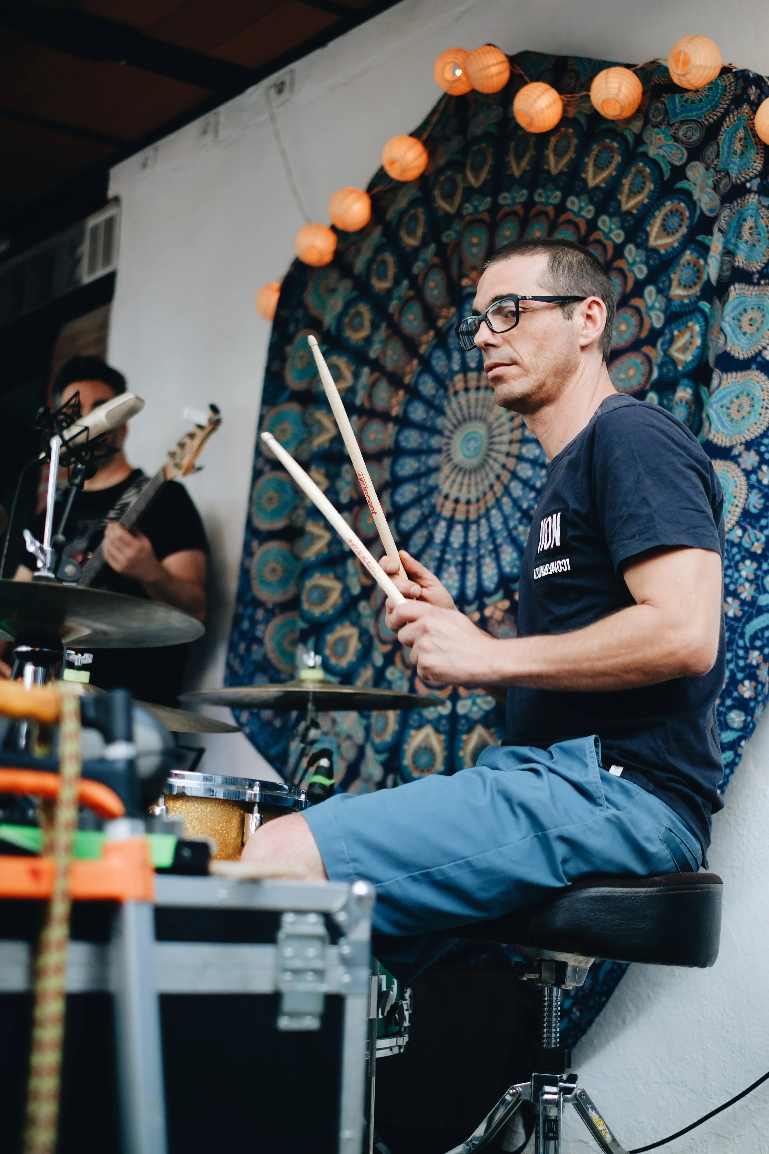 Un hombre tocando la batería en un escenario decorado con mantas y guirnaldas de luces de papel en tonos naranja. En el fondo, hay otra persona tocando la guitarra.