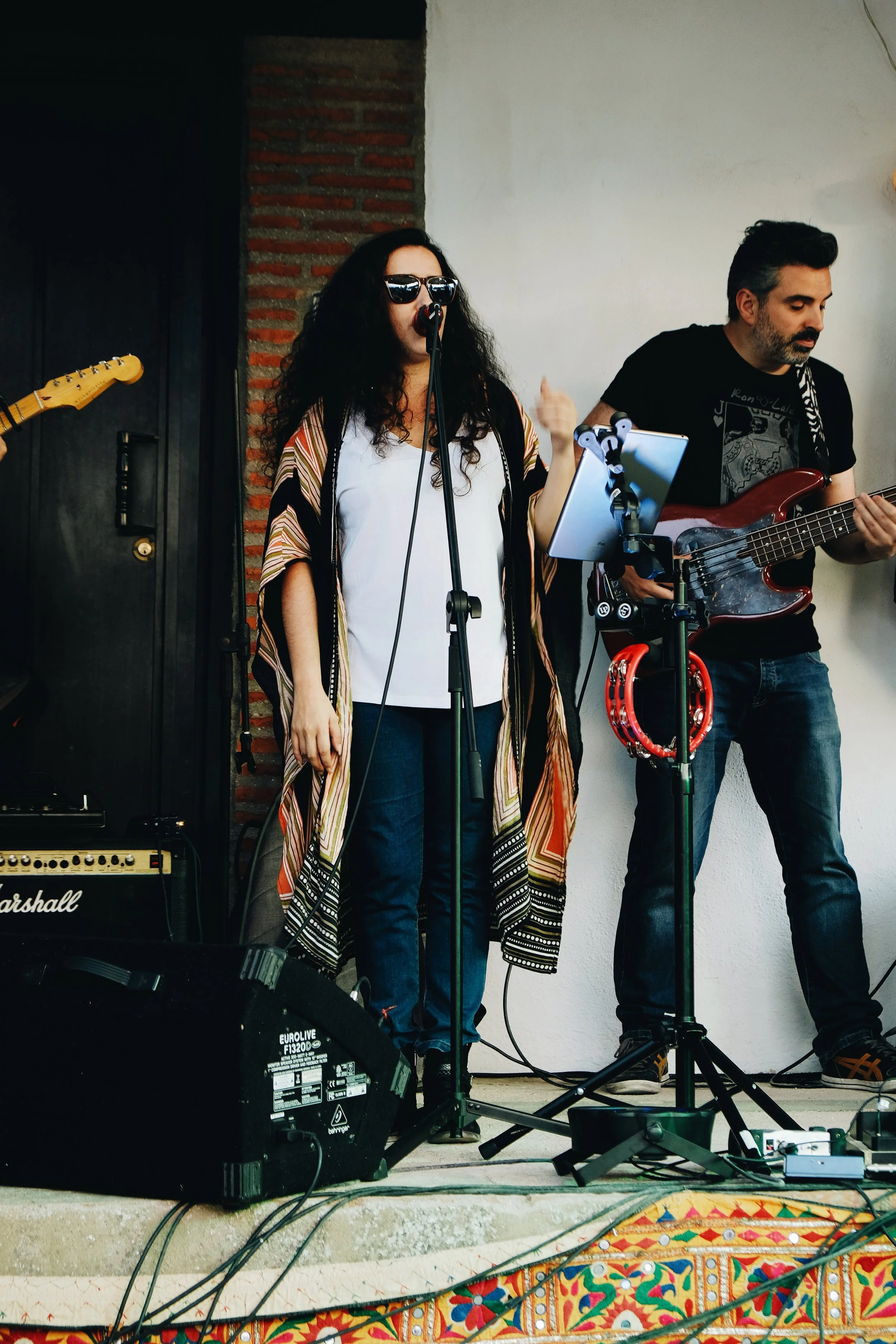 Mujer cantando en un escenario con banda en vivo, tocando la guitarra y en un micrófono, acompañada por un hombre que toca la guitarra eléctrica.