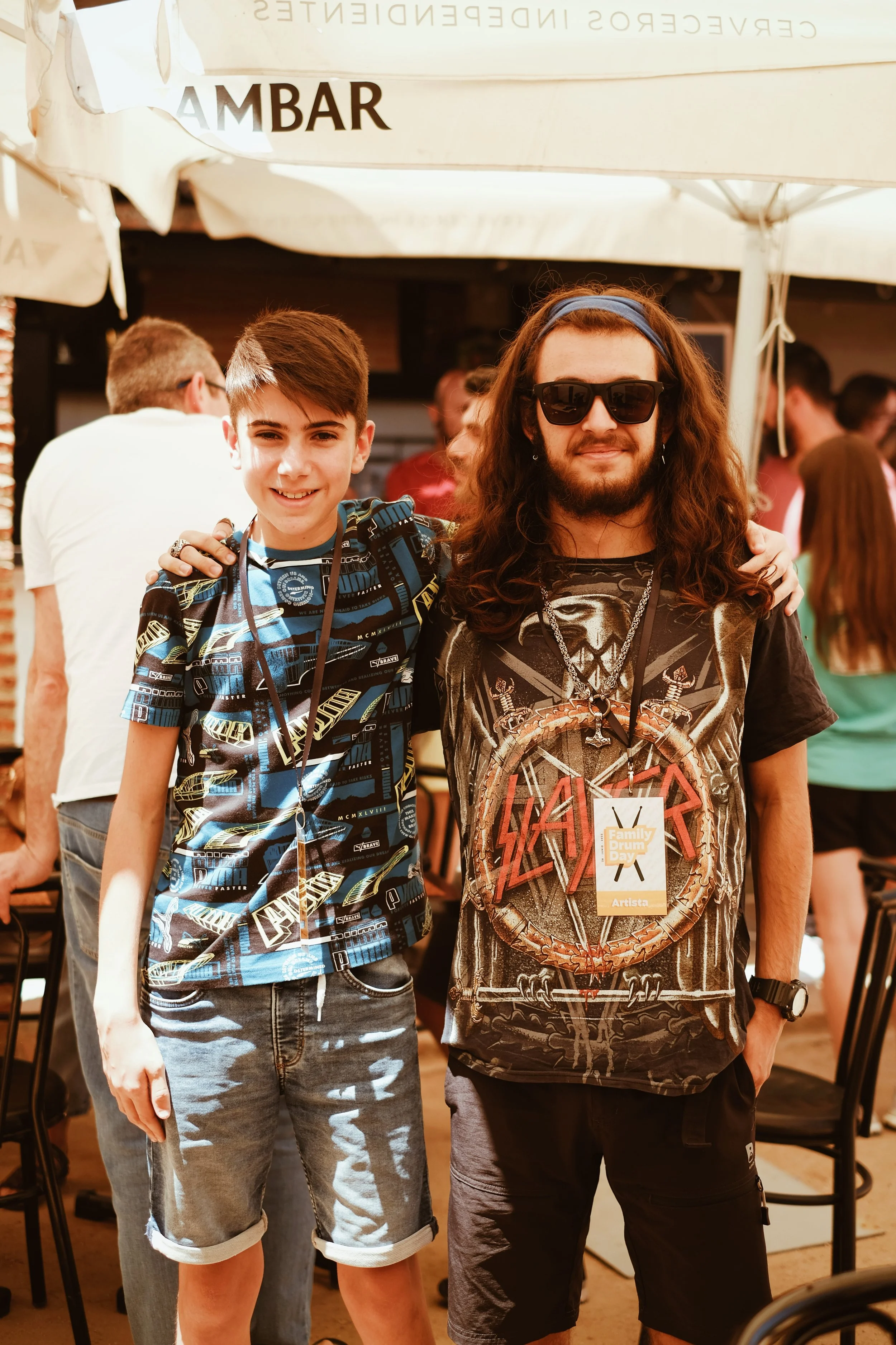 Dos personas con gafas de sol y camisetas de rock, posando en un evento al aire libre bajo sombrillas.