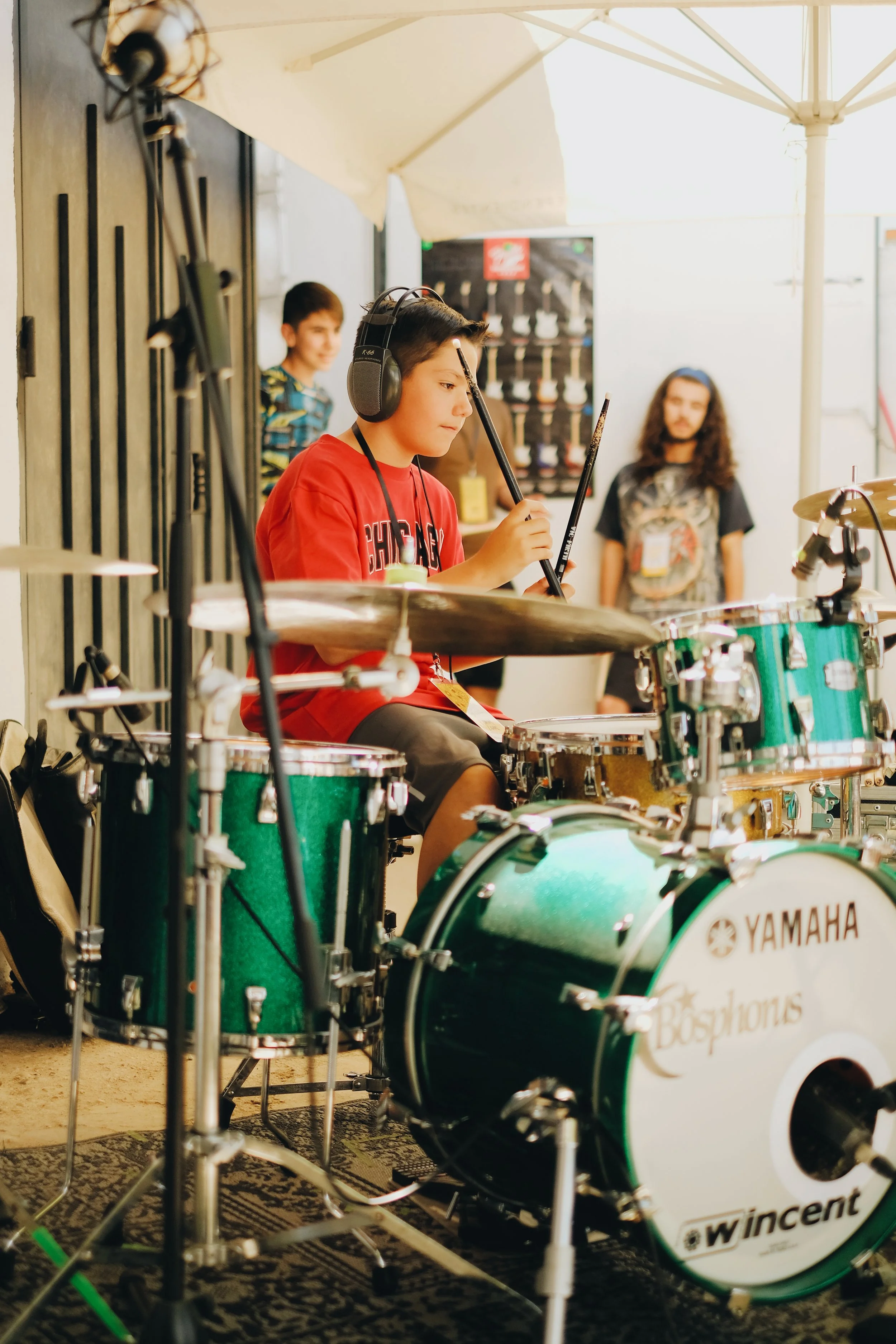 Juvenil tocando batería en un escenario, usando audífonos y concentrado en tocar, con otros niños en el fondo.