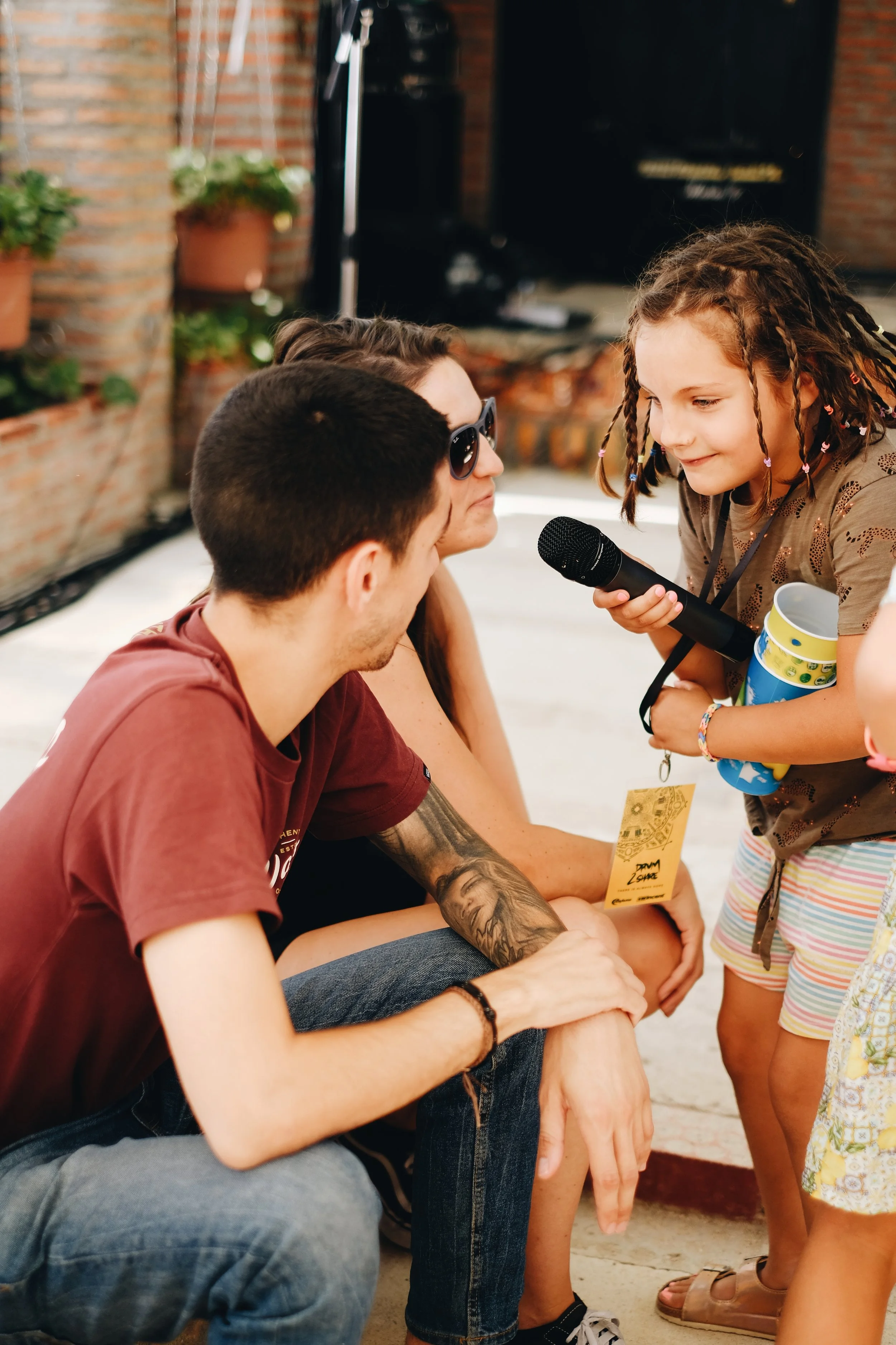 Una niña de cabello rasta y vestimenta casual habla en un micrófono a una pareja de jóvenes sentados, en un ambiente de interior con plantas y ladrillos en el fondo.