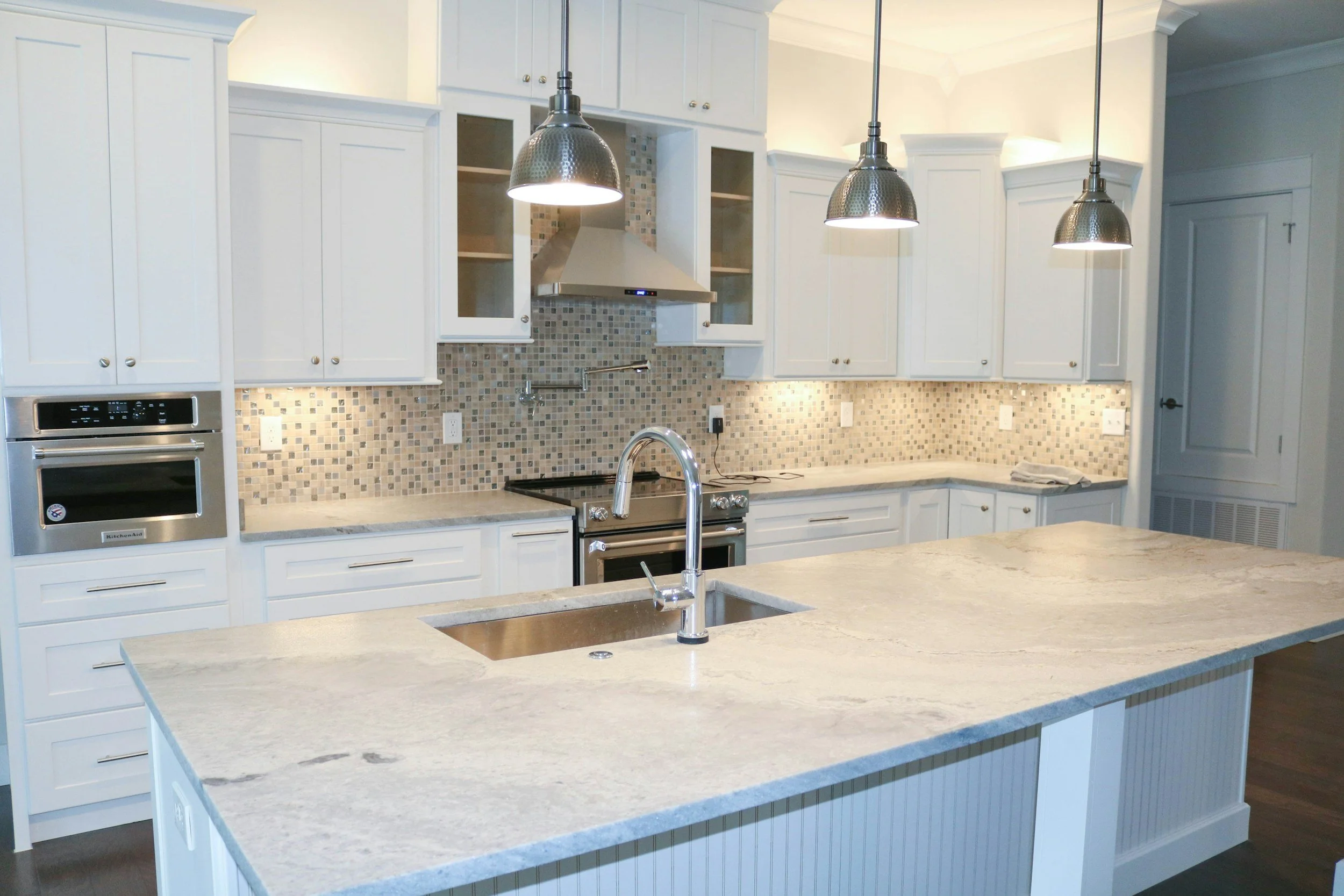 Modern kitchen with white cabinets, a marble island countertop, and stainless steel appliances. Three pendant lights hang over the island, and the backsplash features small mosaic tiles.