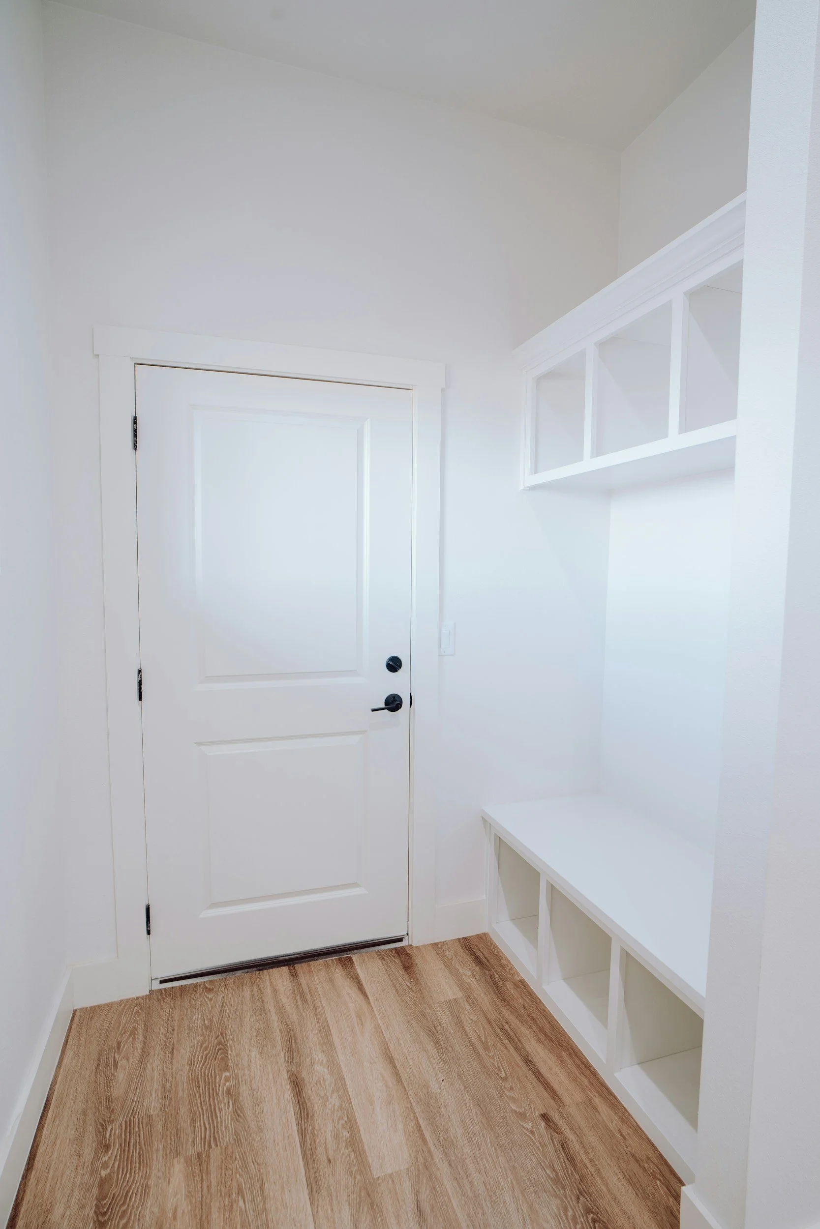 A white interior room featuring a closed white door with black hardware, a built-in white shelf with glass-paneled sections, and a wooden floor.