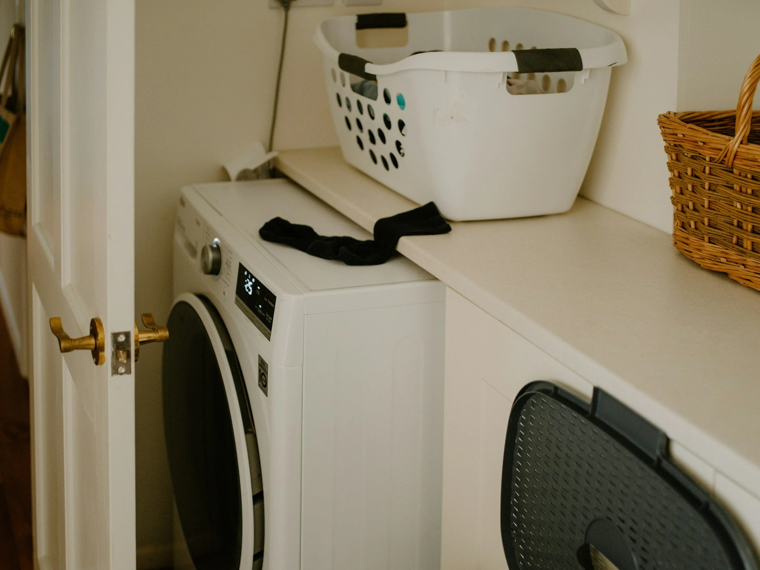 A laundry room with a white washing machine, a white laundry basket on top, a black piece of cloth, a woven basket, and a drying rack.