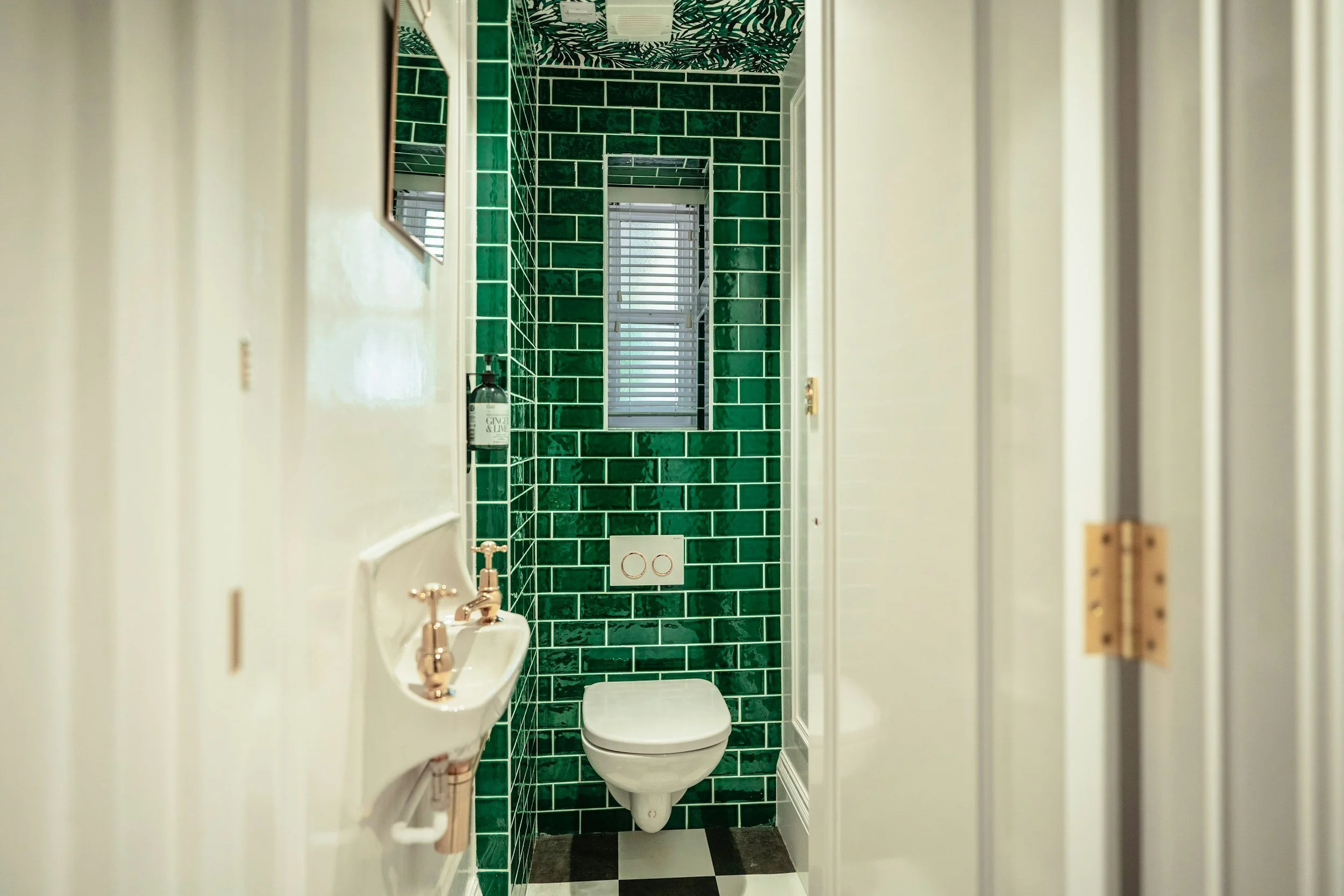 Small bathroom with green tiled wall behind a floating toilet, a narrow window with blinds, a small white sink with gold fixtures on the left, and a black and white checkered floor.