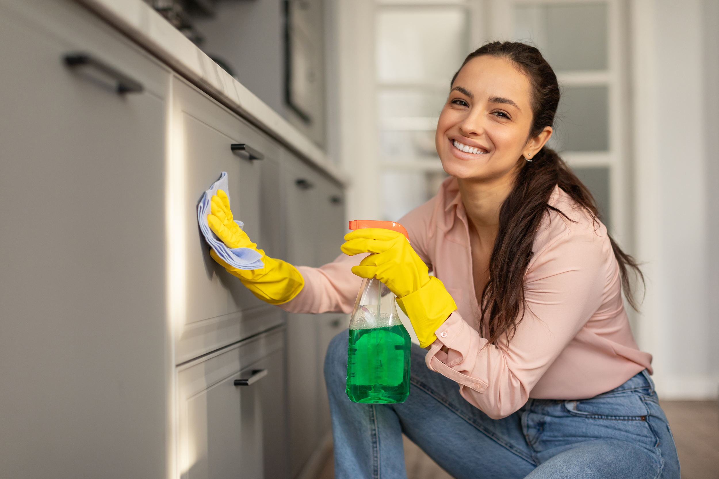 A young woman cleaning a kitchen cabinet with a cloth and spray bottle filled with green cleaning solution, smiling at the camera.