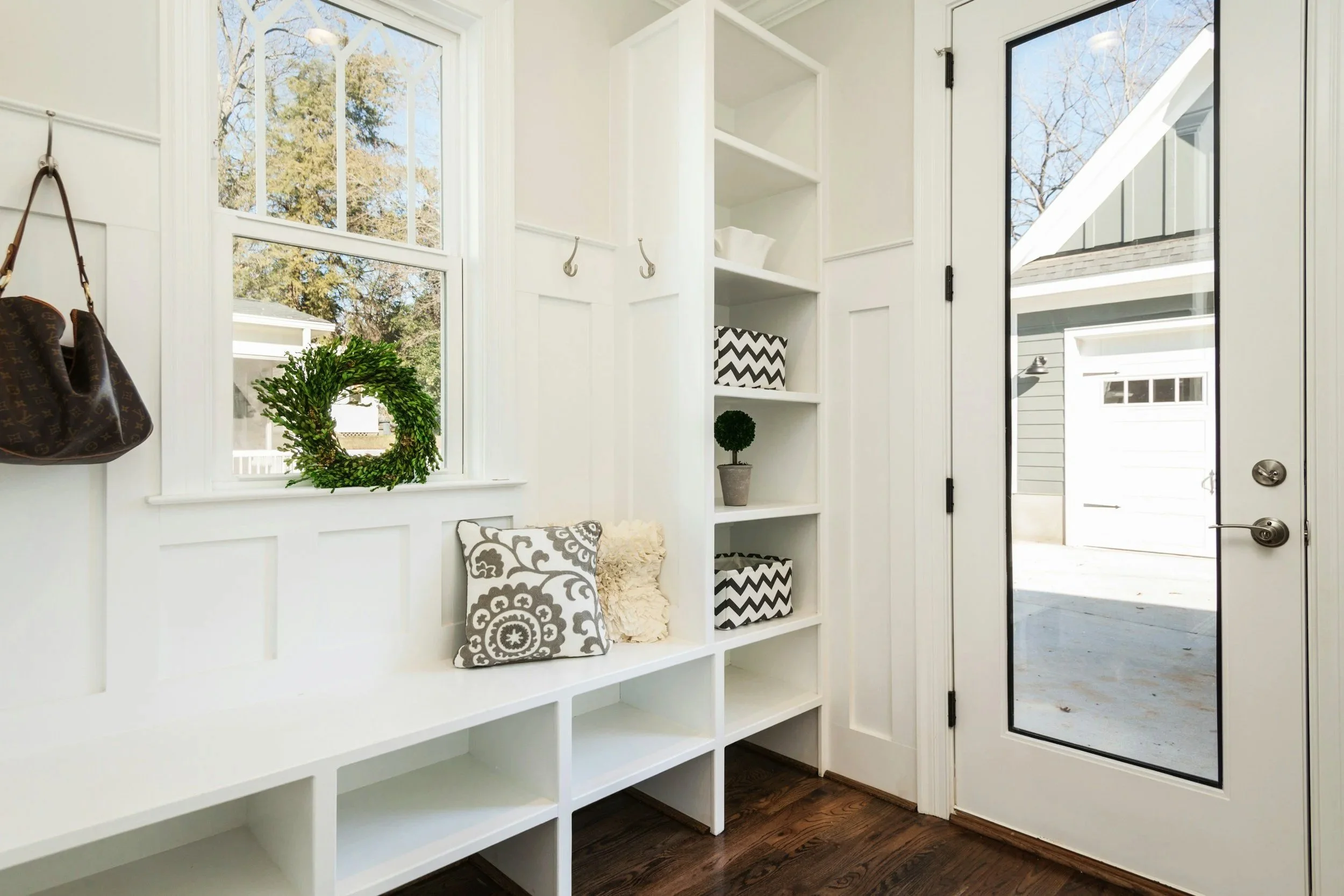 Interior of a white entryway with a bench, decorative pillows, a wall-mounted bag, a window with a wreath, and a door with a mirror showing outside view of a garage and clear sky.