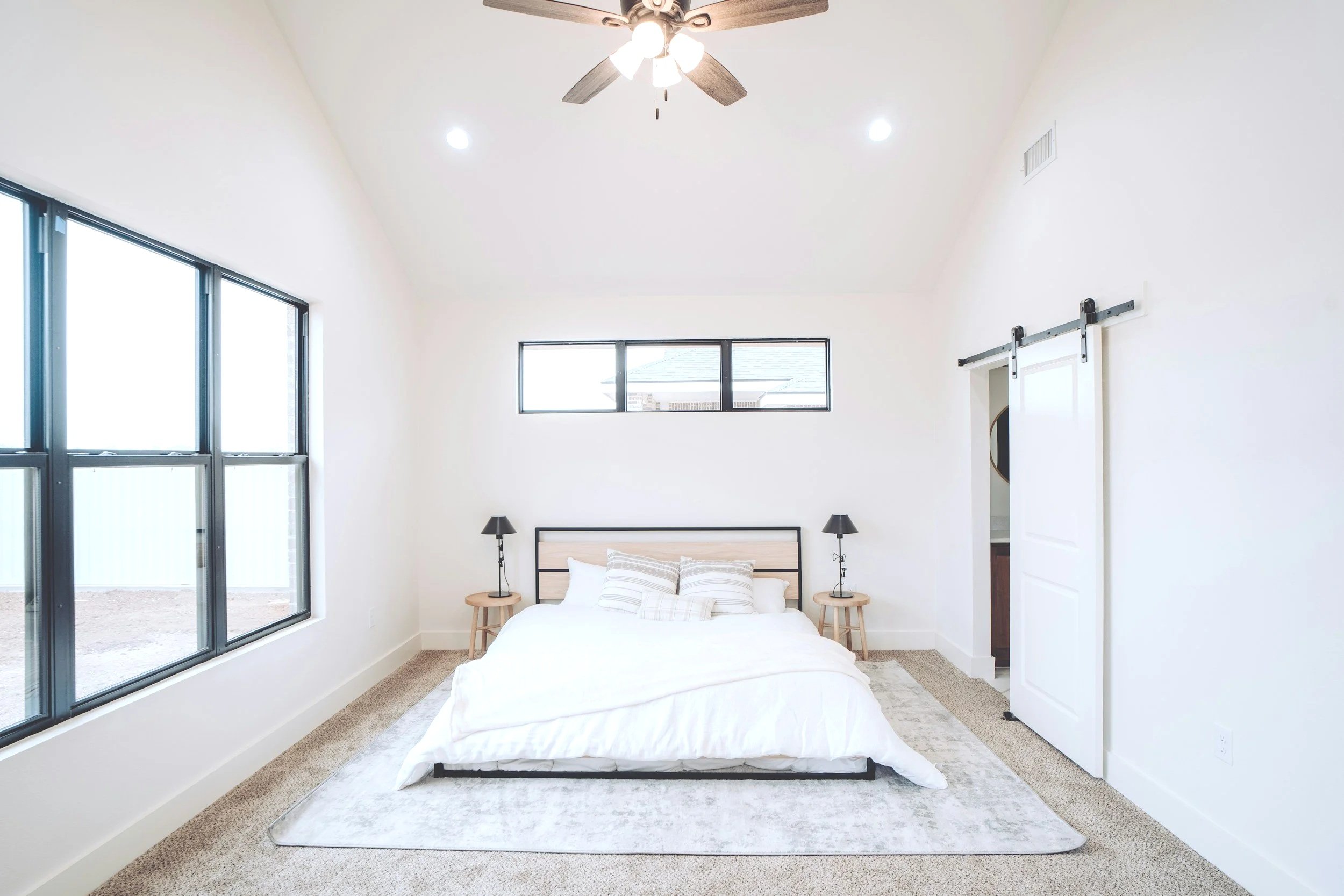 A bright, modern bedroom with a low bed, white bedding, and striped pillows. There are two black bedside lamps on wooden stools, a ceiling fan, large windows on the left side, a small horizontal window above the bed, and a sliding barn door on the right.
